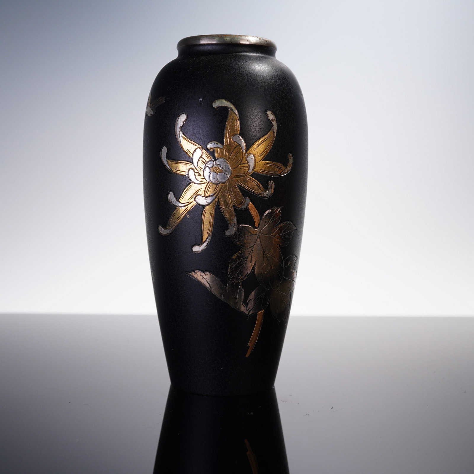 Japanese Mixed Metal Vase with Gilded Floral Motif: A Japanese mixed metal vase, likely late Meiji to early Taisho period, decorated in gold and silver with a chrysanthemum blossom and leafy foliage in relief against a matte black ground. The slender o