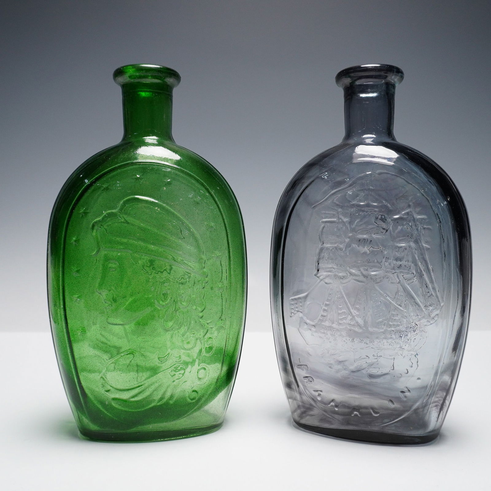 2pc Glass Bottles with Historical Motifs, Green and Gray: Pair of glass bottles, one in olive green depicting a classical profile bust surrounded by stars, and the other in smoky gray featuring a three-masted sailing ship and the word Franklin below. Both fl