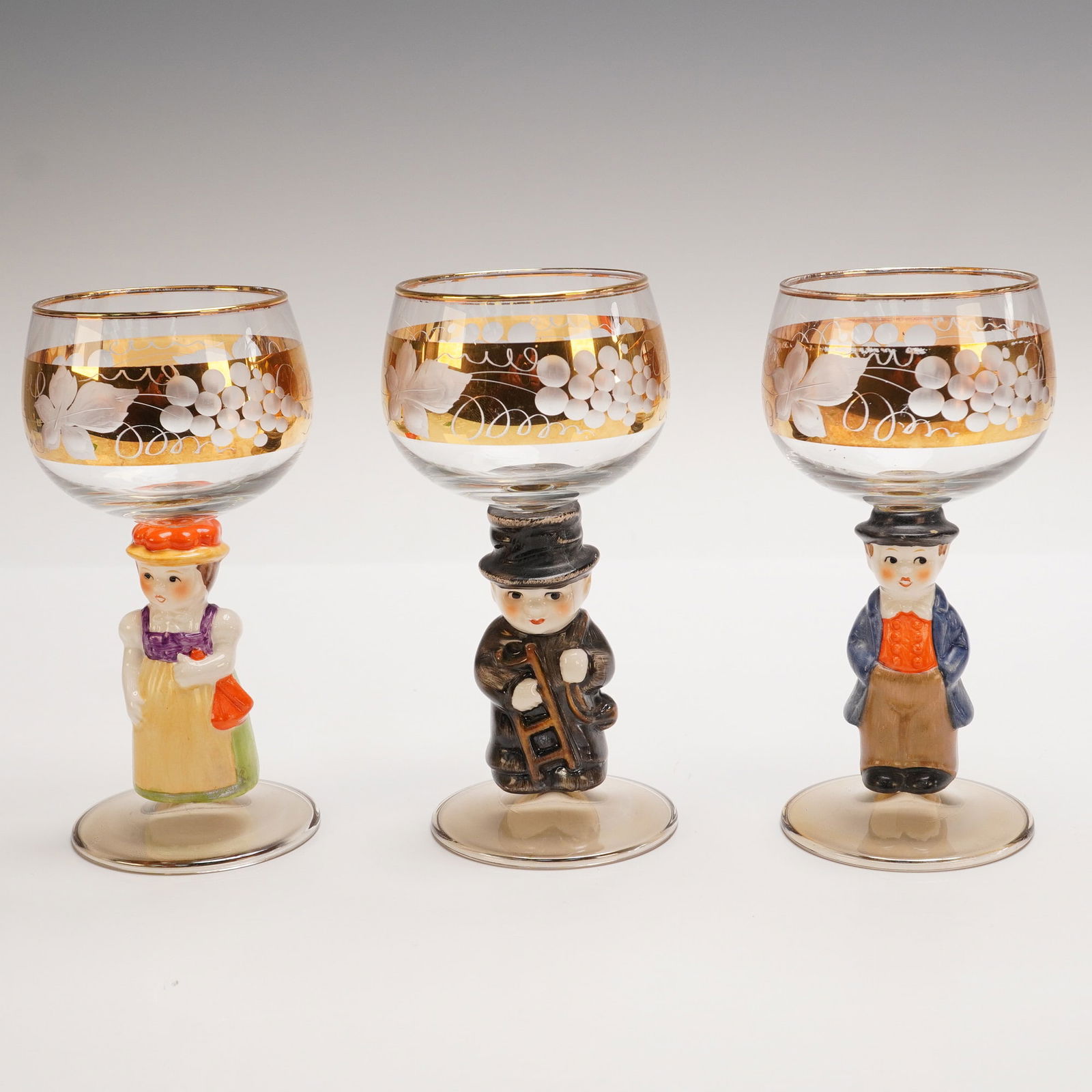 3pc Character Stem Goblets with Gold Floral Etching: Set of three whimsical goblets featuring figural stems of two men and one woman in colorful attire. Each bowl is decorated with an ornate gold band etched with white floral motifs. A charming blend of
