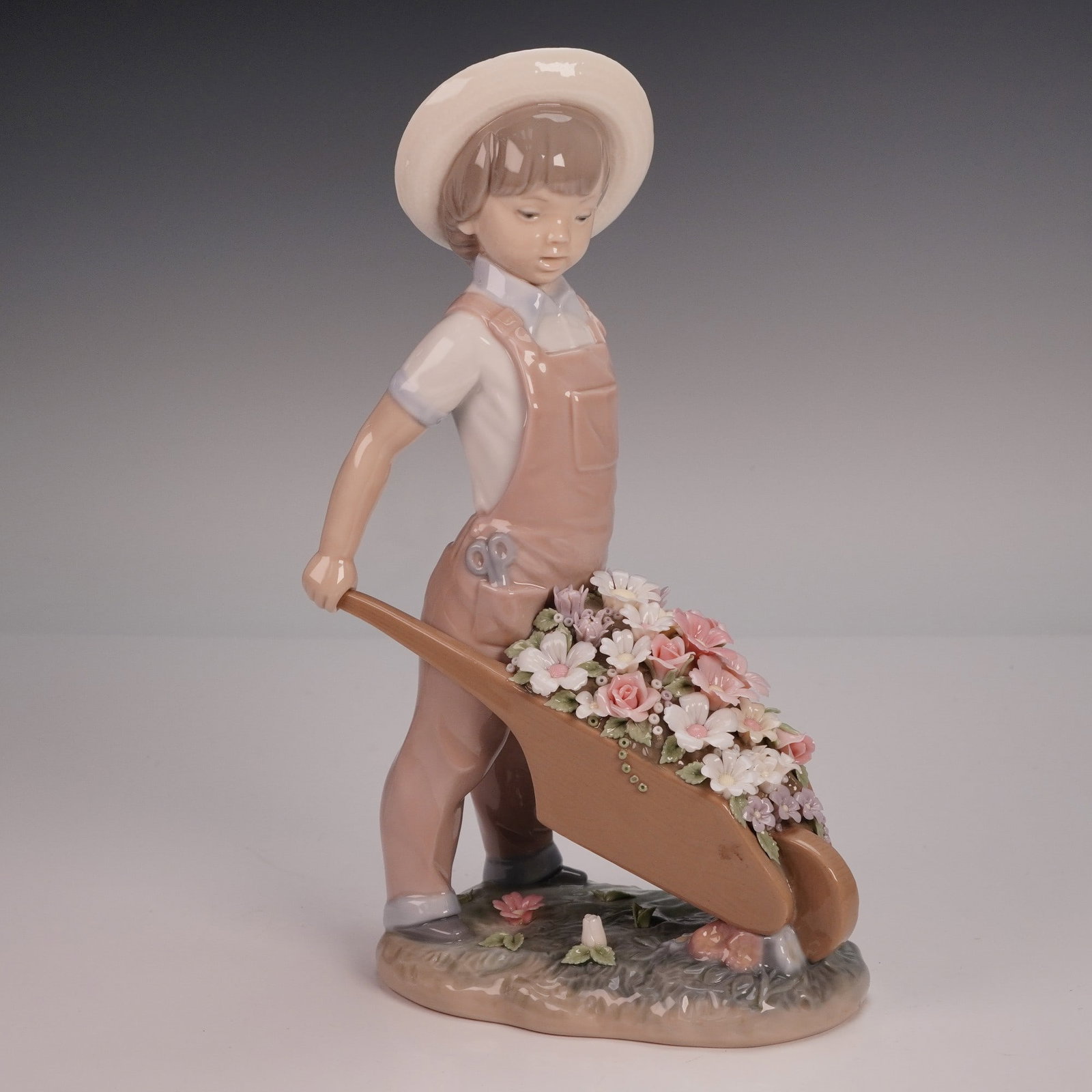 Lladro Porcelain Figurine, Little Gardener 1001283: Porcelain figurine by Lladro titled Little Gardener 1283, sculpted by Francisco Catala. Features a young boy in overalls and a straw hat, wheeling a barrow overflowing with pastel blossoms. Finished i