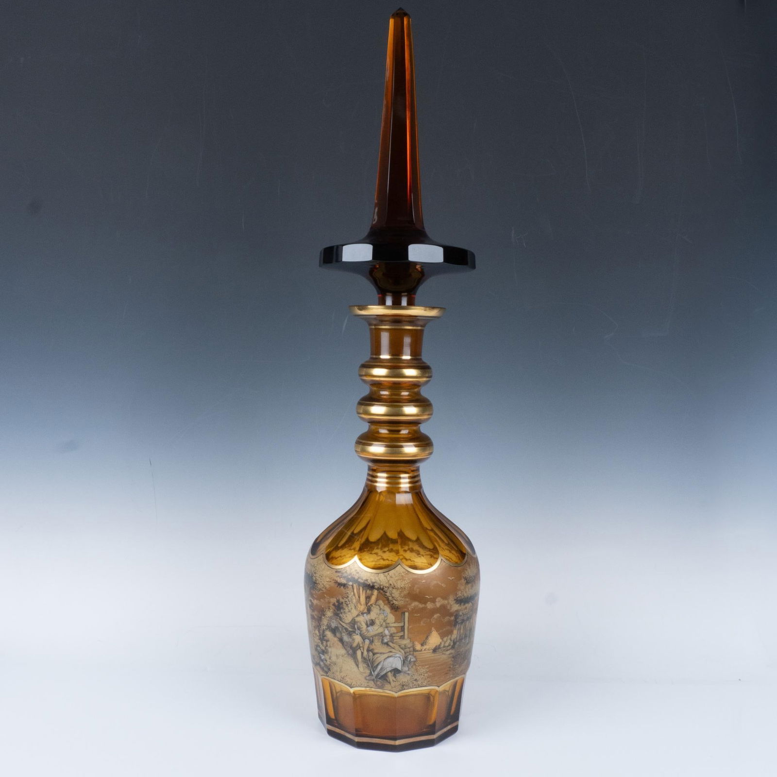 Colossal J.M Pohl Bohemian Czech Glass Large Bottle: Impressive hand blown amber glass vessel featuring an intricately detailed stopper with a sharp, elongated obelisk finial. The body is adorned with an encircling gilt painted Romantic figural scene. T