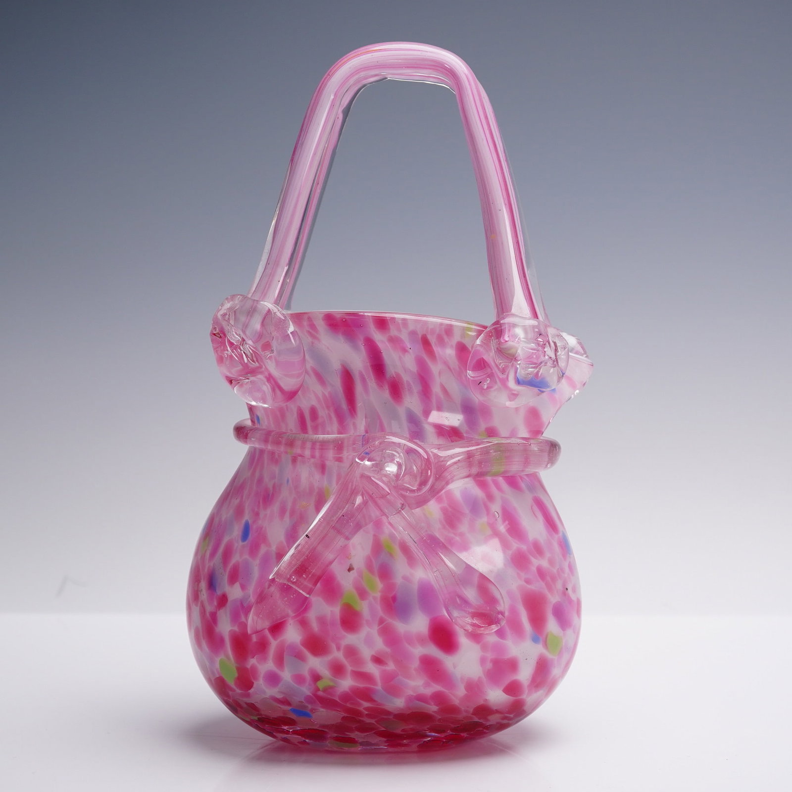 Hand-Blown Art Glass Vase in Pink Confetti with Applied Handle: A vibrant hand-blown art glass vase in the shape of a stylized purse, standing 9 inches tall. This playful, sculptural form features a mottled "confetti" pattern in bright pink with splashes of green,