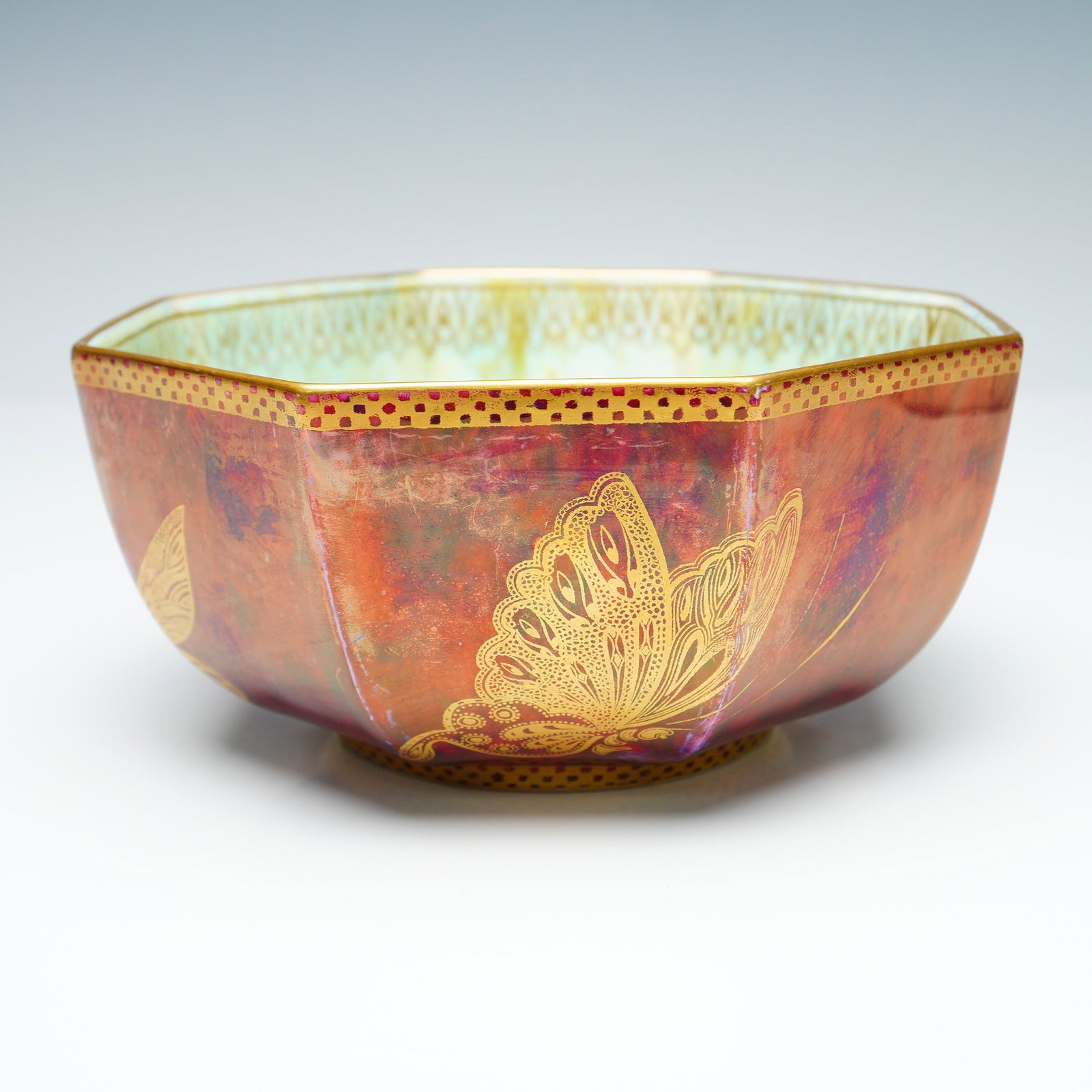 Wedgwood Fairyland Lustre Octagon Bowl, Butterflies Z4827: The exterior is finished in a ruby lustre glaze with subtle mottling in blue tones, further accented with golden butterflies rendered in varying styles. Decorative gold borders with dotted motifs enci