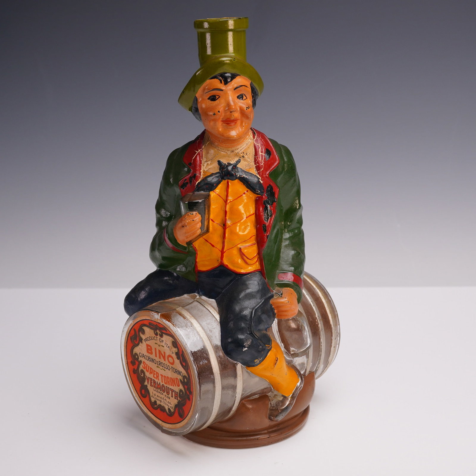 Bino Super Torino Vermouth Figural Bottle: A whimsical figural liquor decanter shaped as a jovial man seated atop a barrel, designed as a promotional bottle for Bino Super Torino Vermouth. The hand-painted figure wears an 18th-century style ou