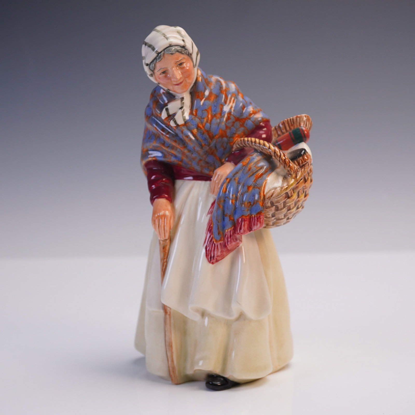 Royal Doulton Figurine, Grandma HN 2052: A heartwarming Royal Doulton figurine titled "Grandma" (HN 2052), portraying a kindly elderly woman in traditional dress, carrying a basket and leaning gently on a cane. Wrapped in a boldly patterned 