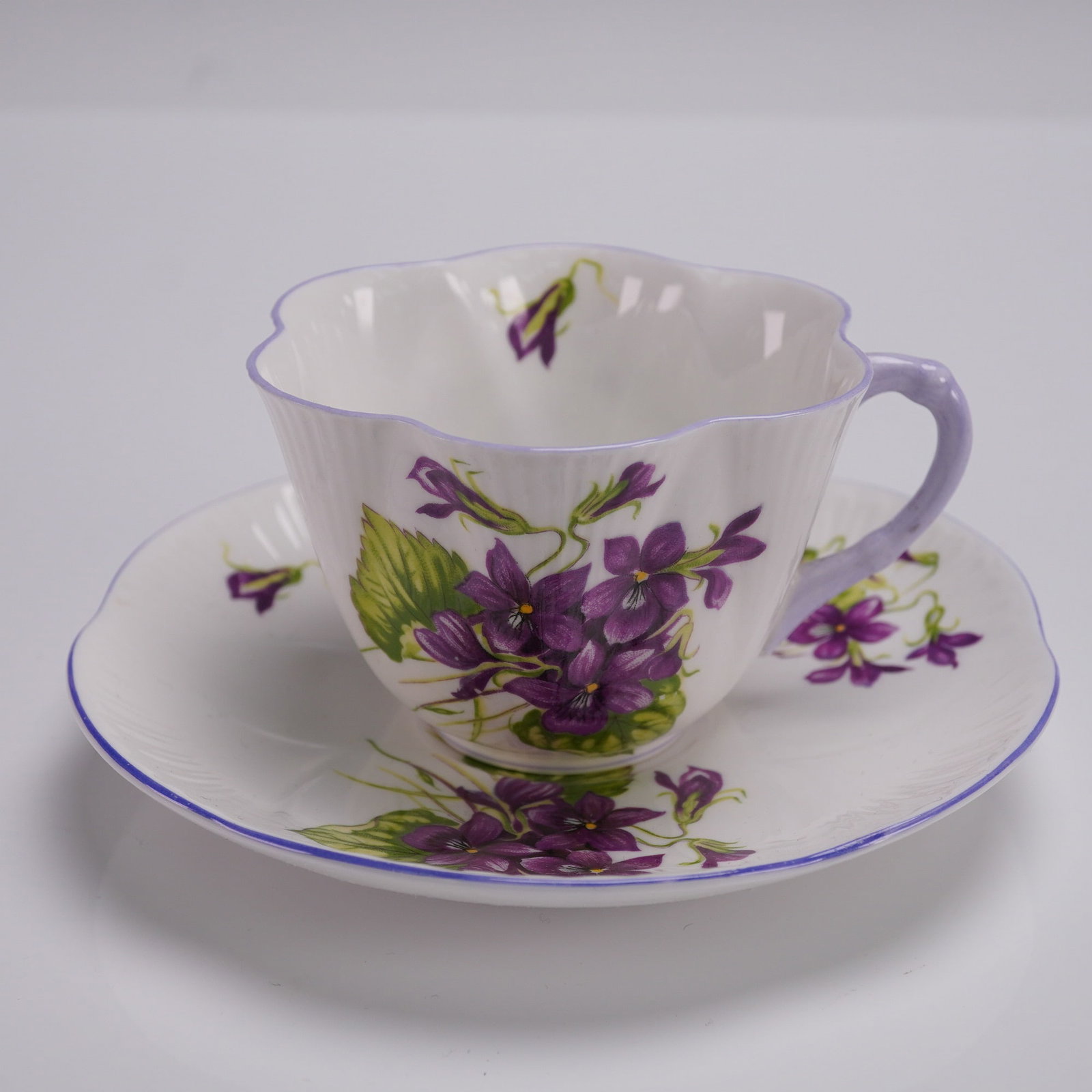 2pc Shelley Fine Bone China Flat Cup and Saucer, Violets Dainty: Shelley fine bone china flat cup and saucer in the Violets pattern, Dainty shape. This elegant 2-piece set features scalloped edges, fluted bodies, and sprays of deep purple violets with green foliage