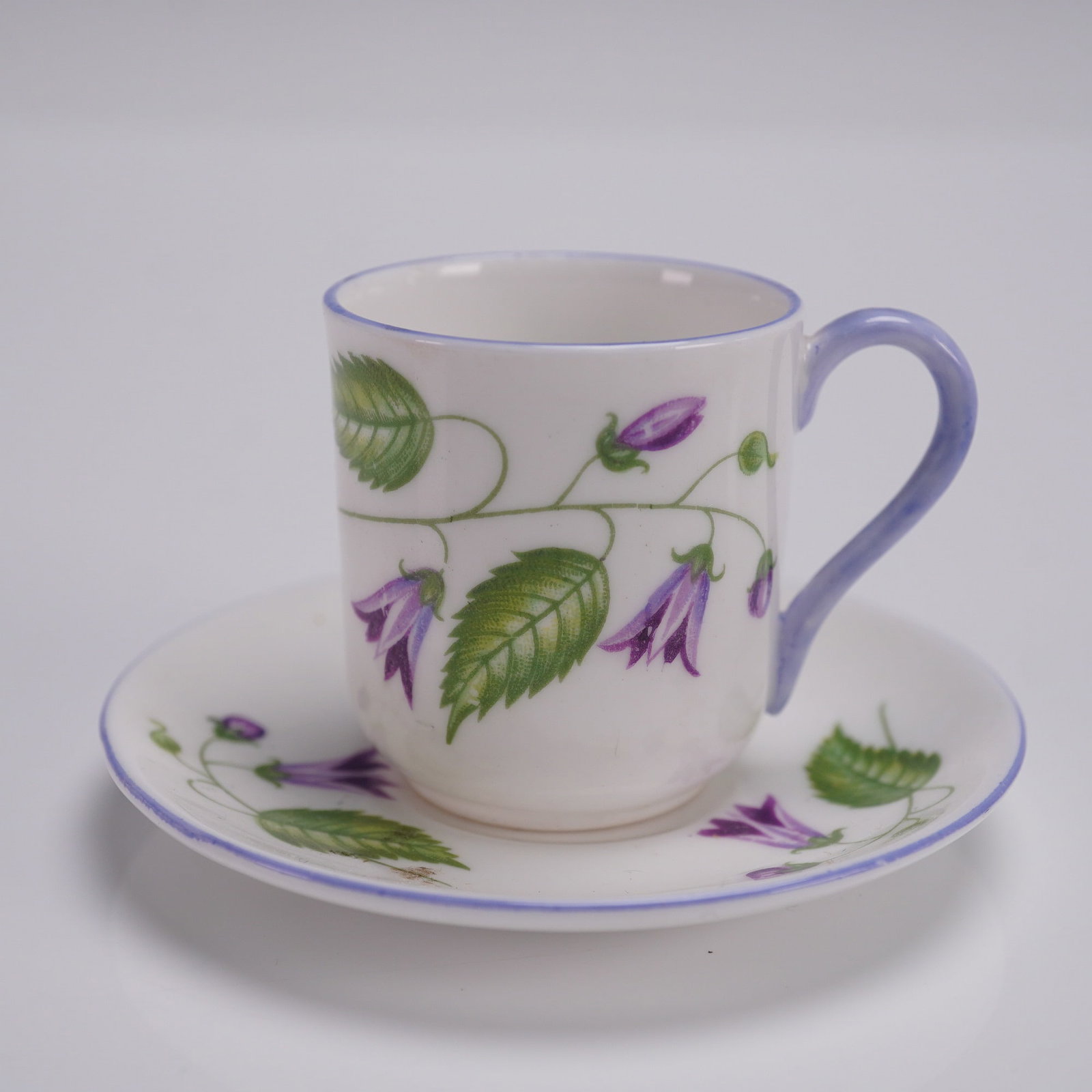 2pc Shelley Miniature Demitasse Cup and Saucer, Campanula: A petite Shelley fine bone china demitasse cup and saucer set in the Campanula pattern, featuring vibrant violet bellflowers and serrated green leaves on a trailing vine. The cylindrical cup and match