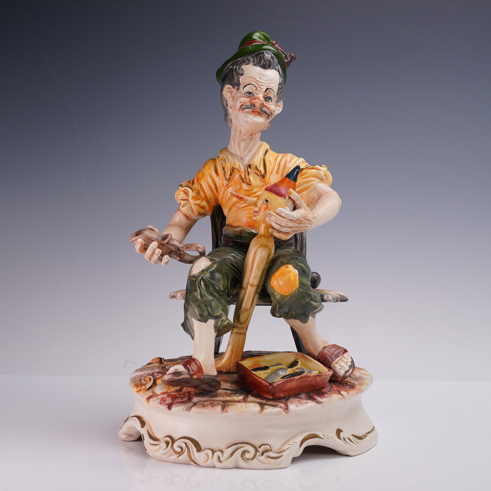 Capodimonte Porcelain Figurine, Cobbler at Work: Hand-painted Capodimonte porcelain figurine depicting a cobbler seated on a stool as he repairs a wooden toy puppet, dressed in a yellow shirt, green trousers, and sandals, with a small toolbox at his