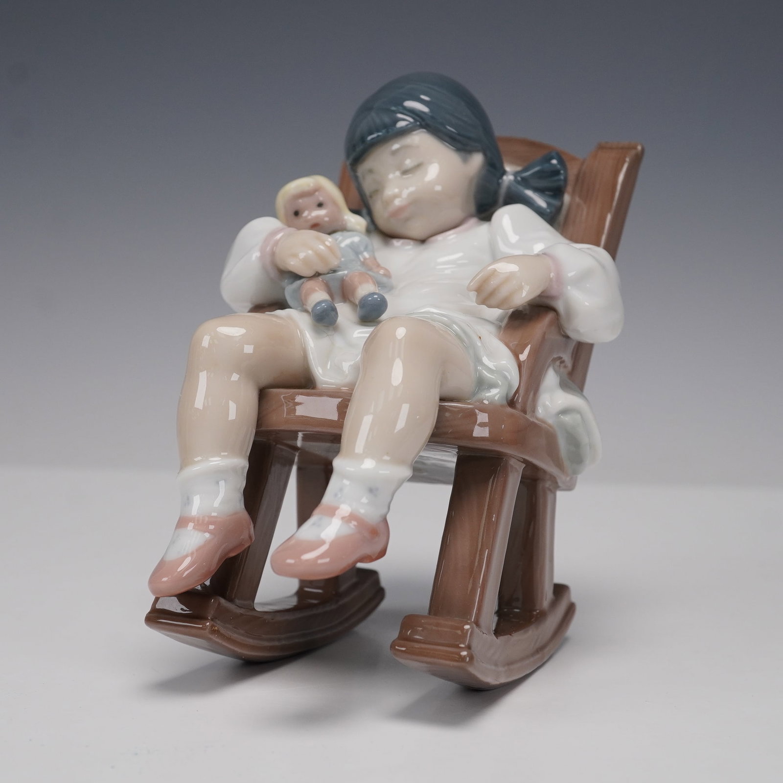Lladro Porcelain Figurine, Naptime 1005448: Glossy porcelain figurine by Lladro titled Naptime, model number 1005448, sculpted by Jose Puche. Features a young girl asleep in a wooden rocking chair, clutching her doll. Issued in 1987 and retired