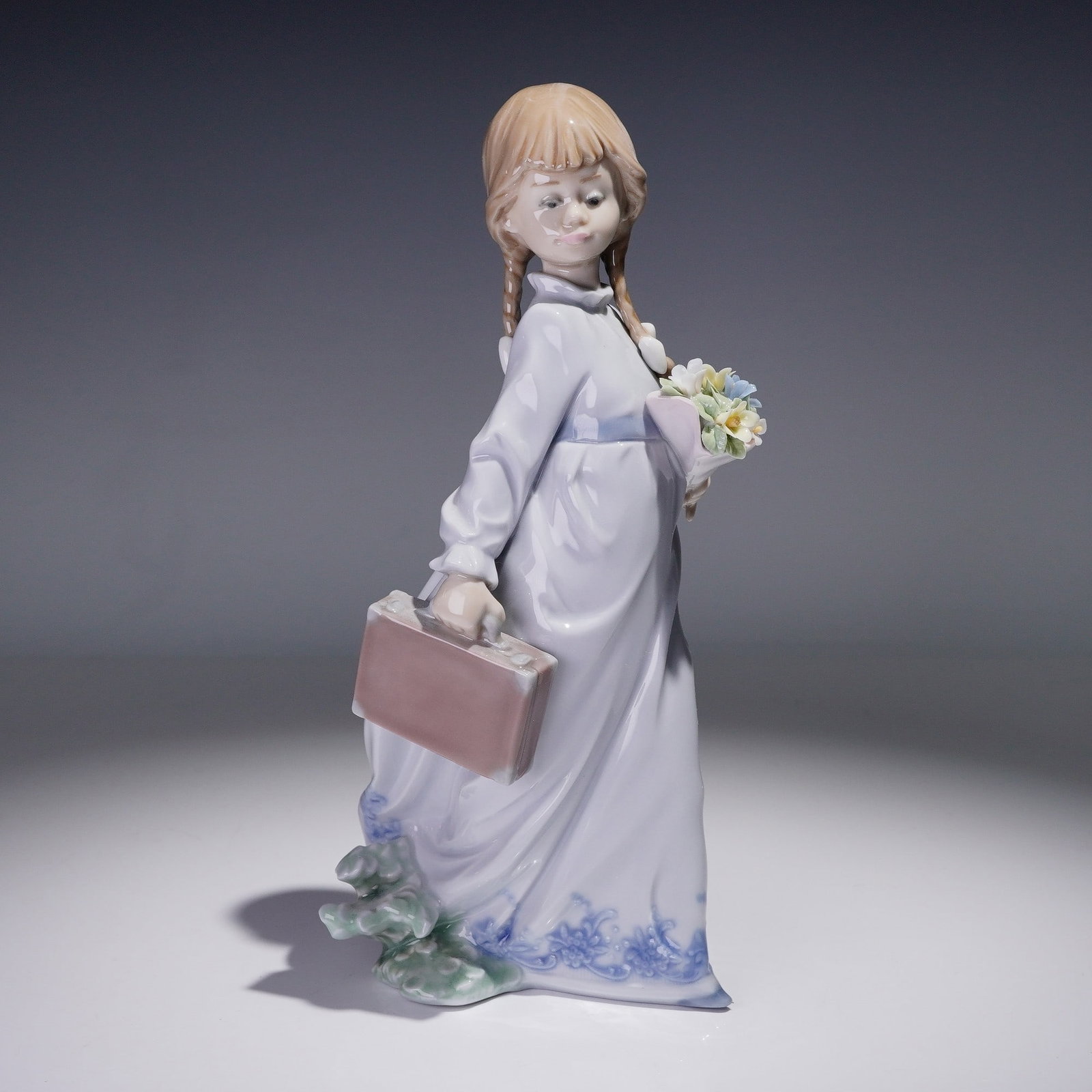 Lladro Porcelain Figurine, School Days 1007604: This glazed porcelain figure depicts a young girl with braided hair, carrying a bouquet of pastel-colored flowers in one hand and a school satchel in the other. She wears a long, flowing dress accente