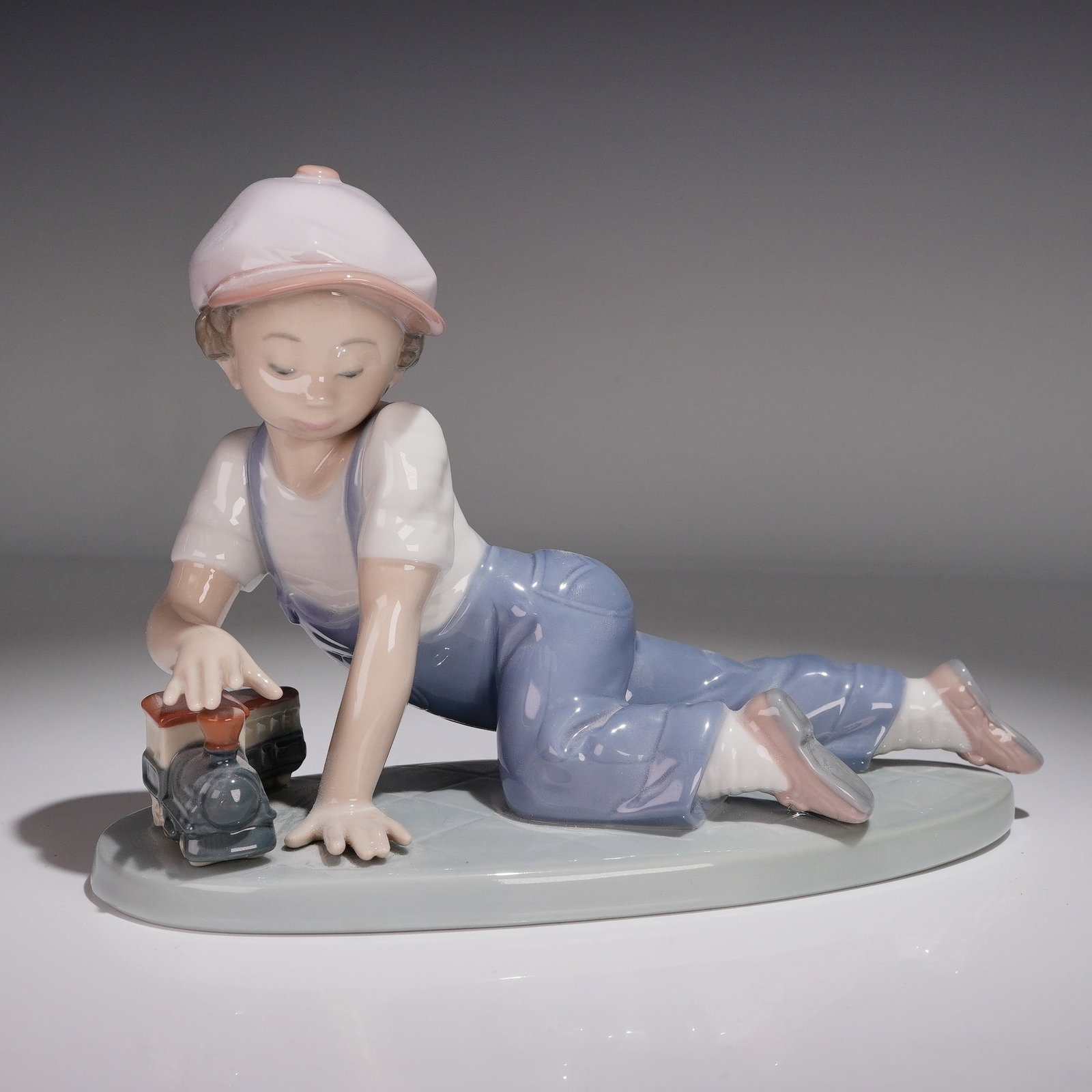 Lladro Porcelain Figurine. All Aboard 1007619: Glossy finish figurine modeled as a young boy in blue overalls playing with a toy train. Lladro backstamp. Artist: Antonio Ramos Issued: 1992 - 1995 Dimensions: 3"L x 7.25"W x 5"H Country of Origin: S