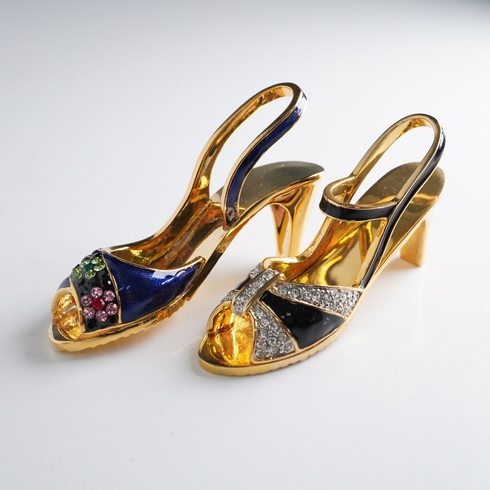 Pair of Rucinni Jeweled Enameled High Heel Figurines: A dazzling pair of miniature high heel shoe figurines by Rucinni, each rendered in rich enamel and sparkling rhinestones. One shoe features a multicolored floral motif on a dark blue ground, while the