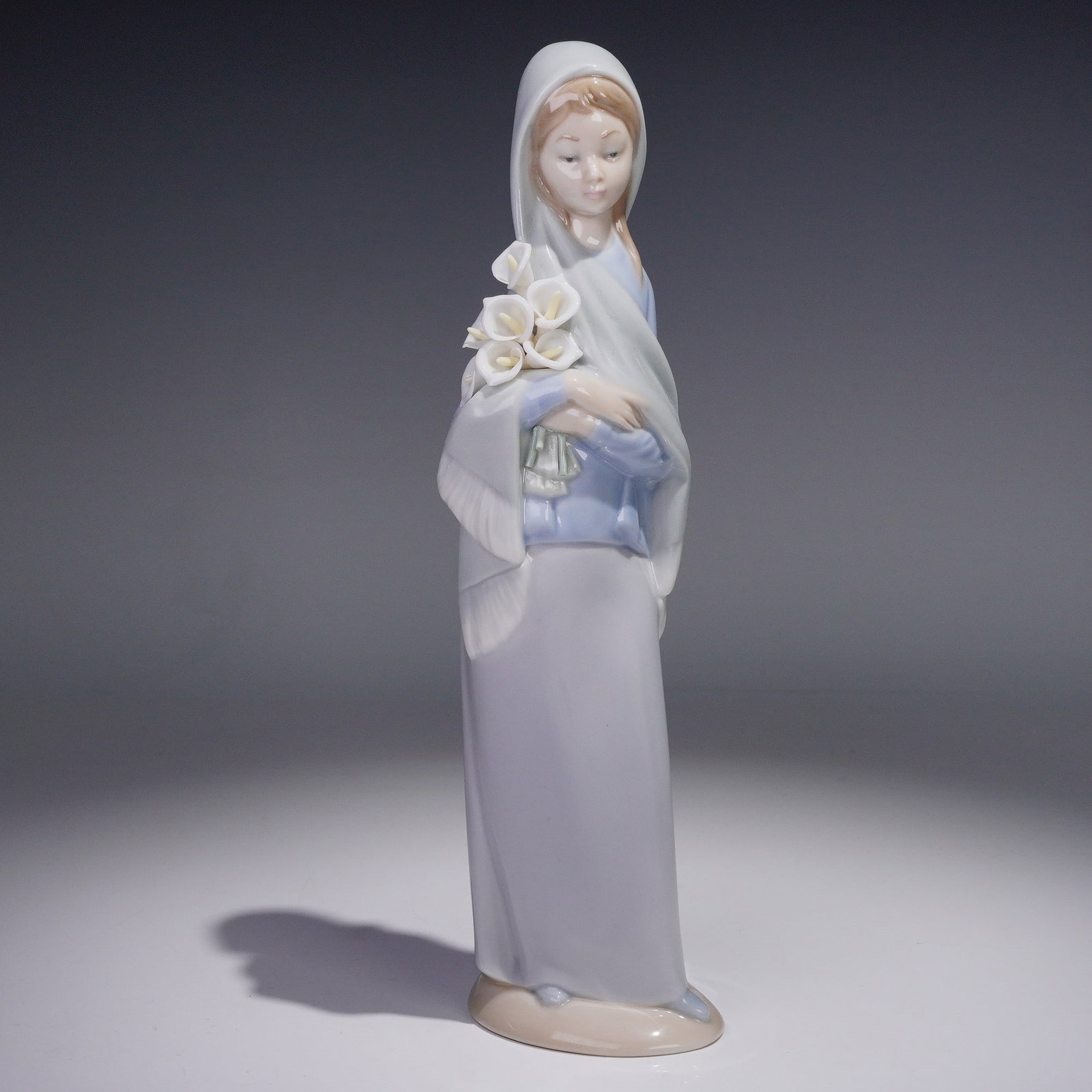 Lladro Porcelain Figurine, Girl with Calla Lilies 1004650: Glossy finish porcelain modeled as a young lady in a cornflower-blue shawl cradling white flowers in her arms. Lladro backstamp. This item has its original box. Box measures: 11"L x 5.5"W x 5"H. Artis
