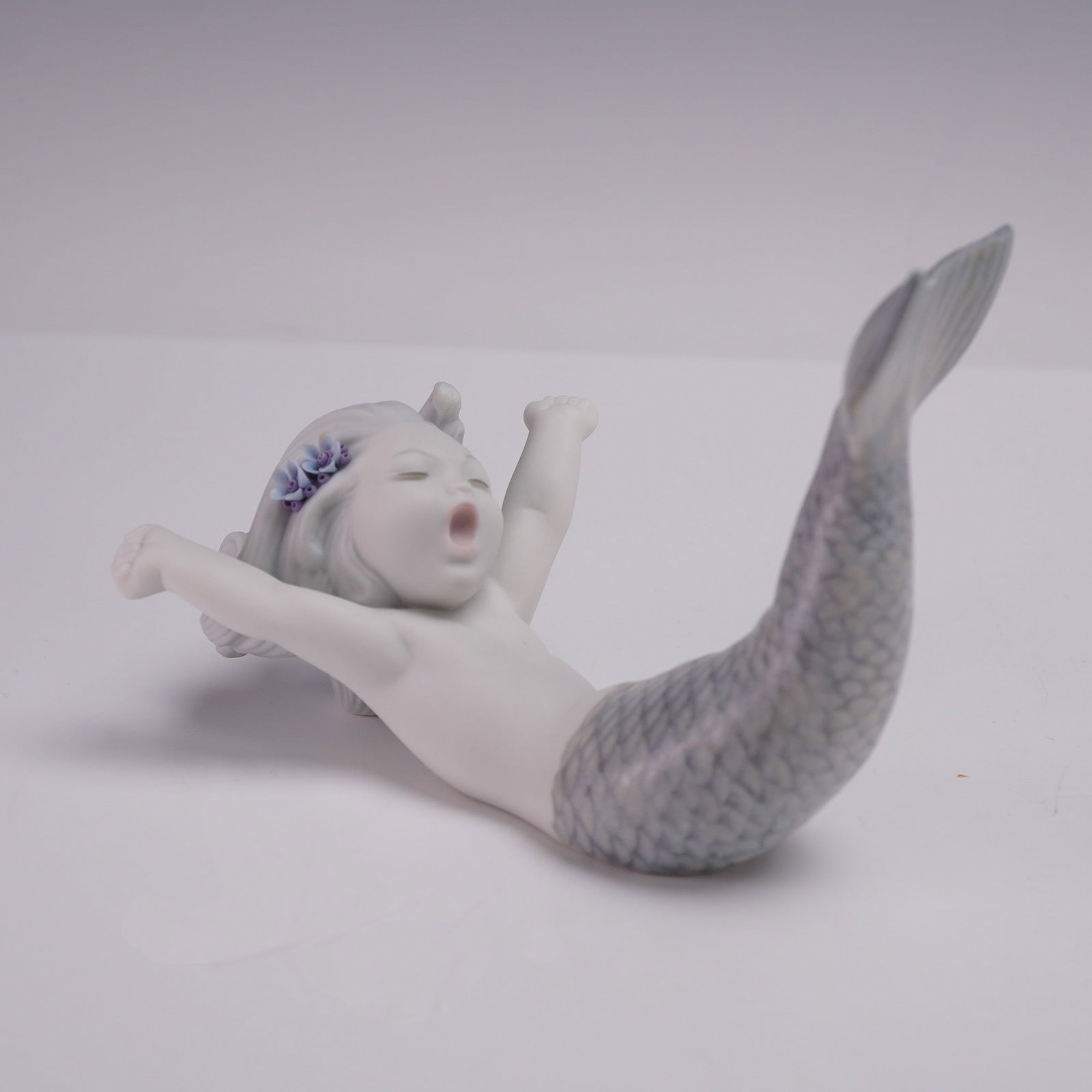 Lladro Porcelain Figurine, Waking Up At Sea 1018113: The matte-finish sculpture depicts a young mermaid stretching with arms extended and mouth open, as if yawning. Features soft pastel tones with detailed hand-painted blue and lavender floral accents i