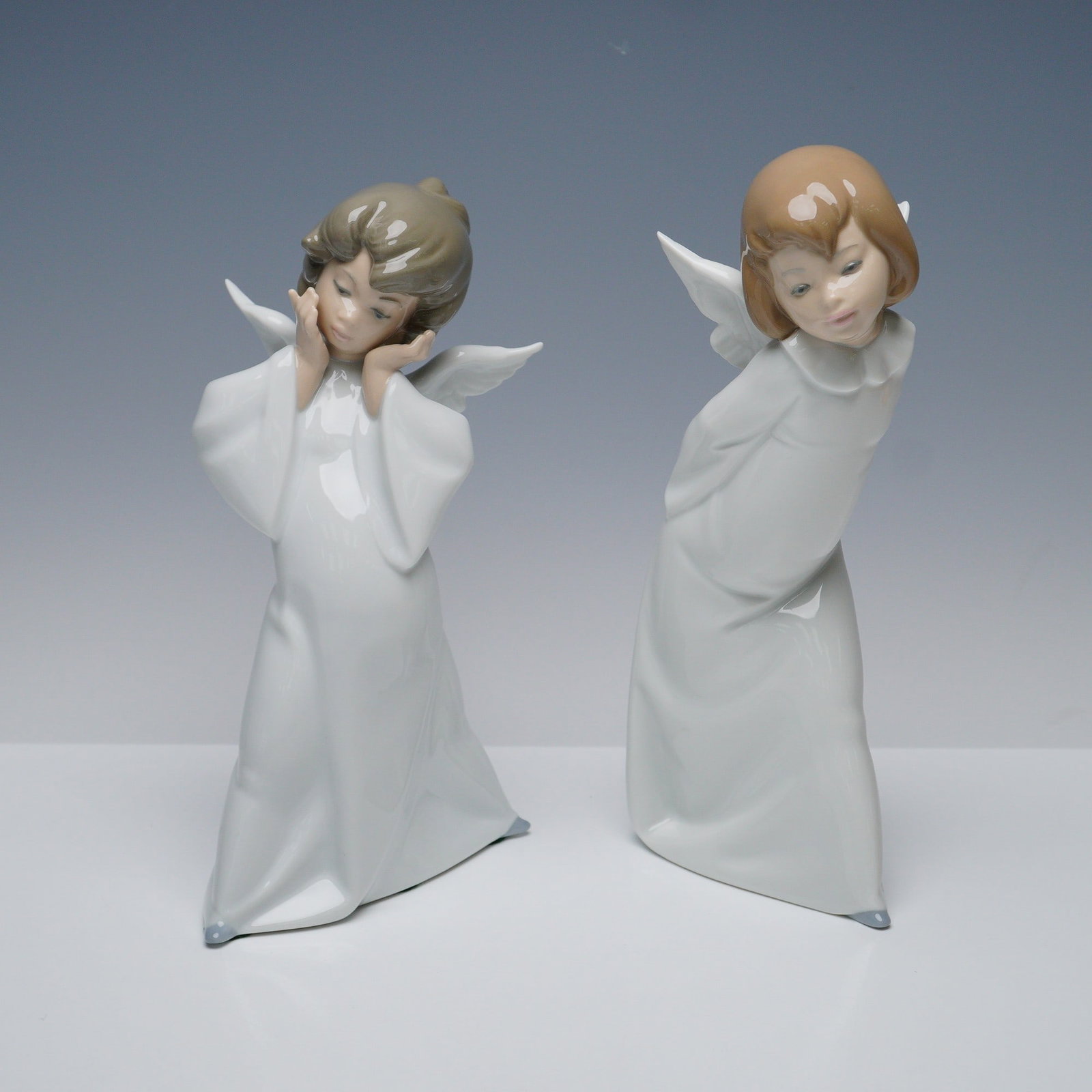 Pair of Lladro Figurines, Cherub Puzzled and Cherub Smiling: Pair of hand-painted porcelain cherub figurines. The pair depicts a cherub with hands held to the face in a puzzled expression (1004959), and a smiling cherub leaning forward while hiding a lantern be