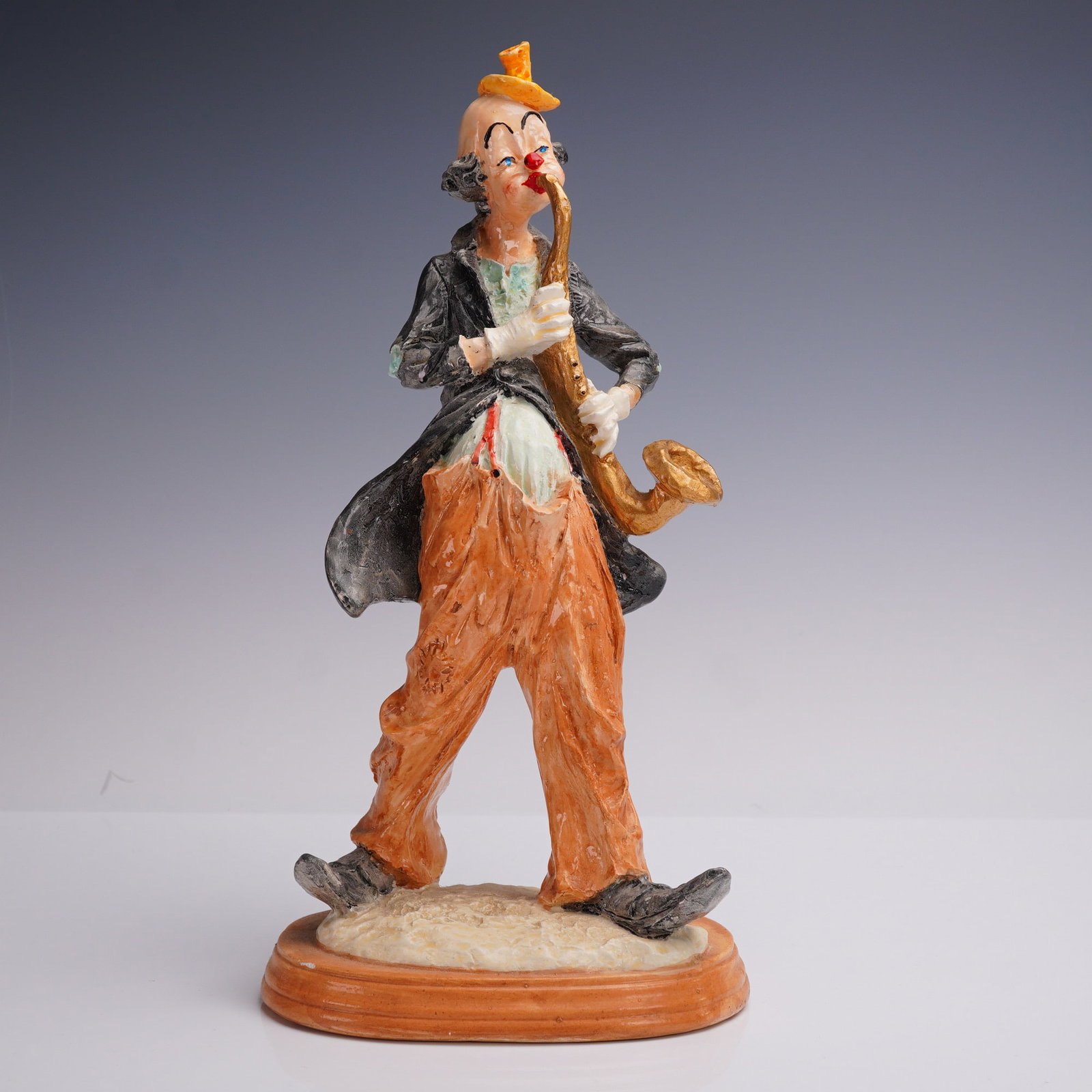 Whimsical Porcelain Clown Figurine with Saxophone: A hand-painted porcelain clown figurine modeled in a humorous stance playing a saxophone. The clown is dressed in exaggerated orange trousers, a black tailcoat, and yellow hat, with expressive facial 