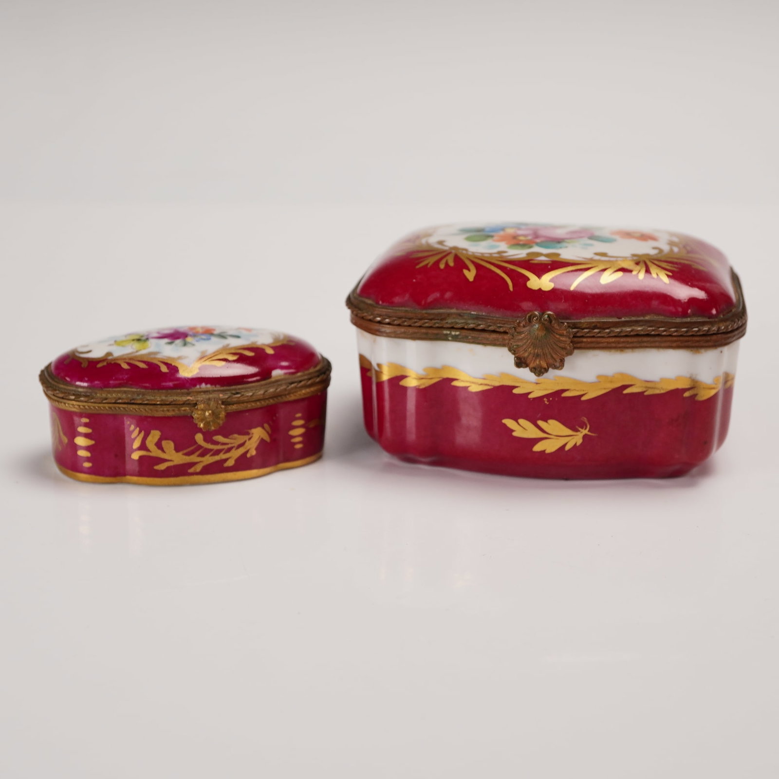 2pc Limoges Hand Painted Floral Decorative Lidded Boxes: A refined pair of Limoges lidded boxes, hand painted with a dark pink exterior. The coloration, surrounded by floral motifs in the center, gilt finished throughout. The interior of the large box is de