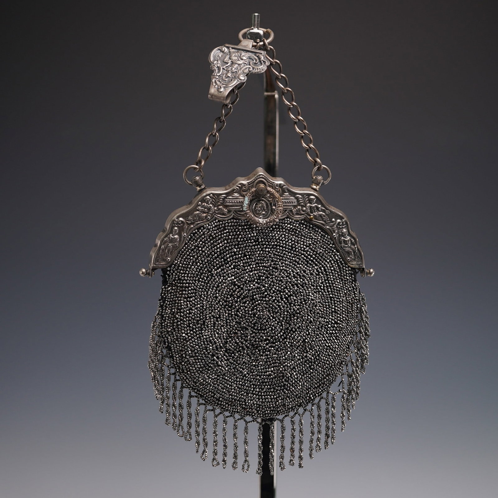 Victorian Style Silver Toned Beaded Handbag: Striking circular evening handbag featuring silver toned beads stitched in tight concentric rings, finished with a skirt of beaded tassels. The silver toned frame is richly decorated with scenes of se