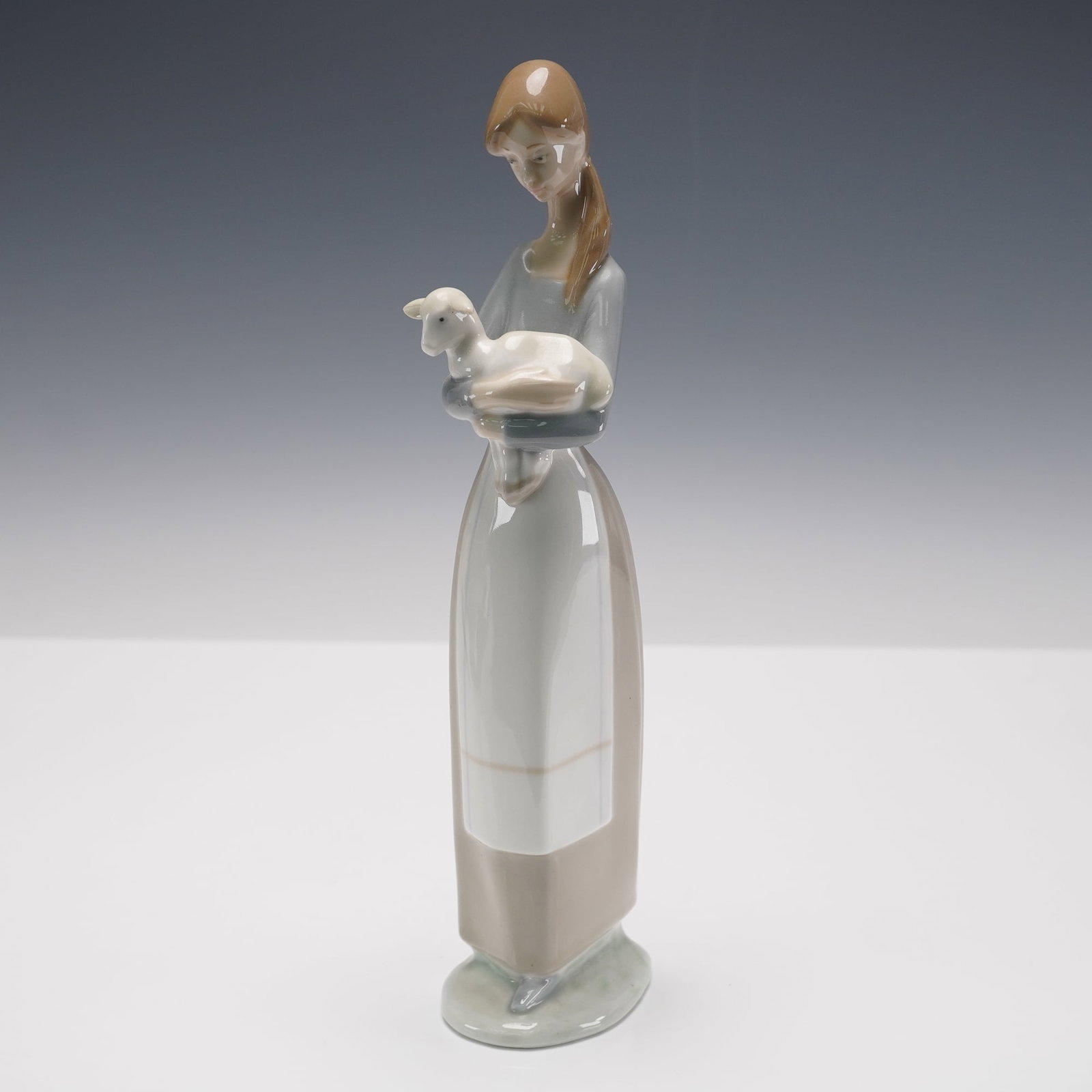 Lladro Porcelain Figurine Girl with Lamb 01004505: Lladro porcelain figurine titled Girl with Lamb, item number 01004505, sculpted by Fulgencio Garcia. This serene and slender figure features a young woman holding a resting lamb in her arms. She wears
