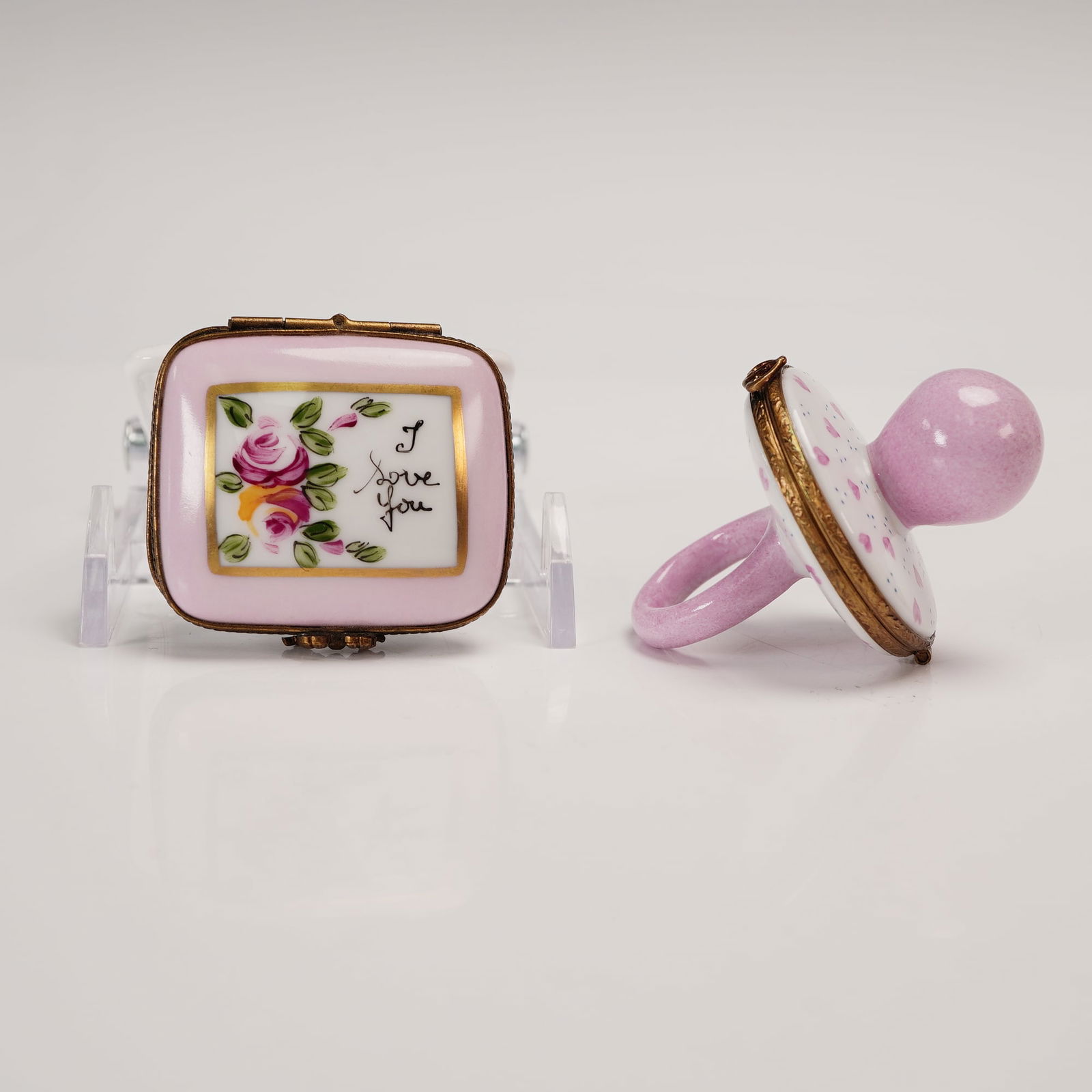 2pc French Limoges Hand Painted Decorative Lidded Boxes: A finely pair of Limoges decorative boxes, including a pink pacifier box designed in a limited edition 11 of 500, and a pink square box. The pacifier is hand painted in hues of pink and blue specks. T