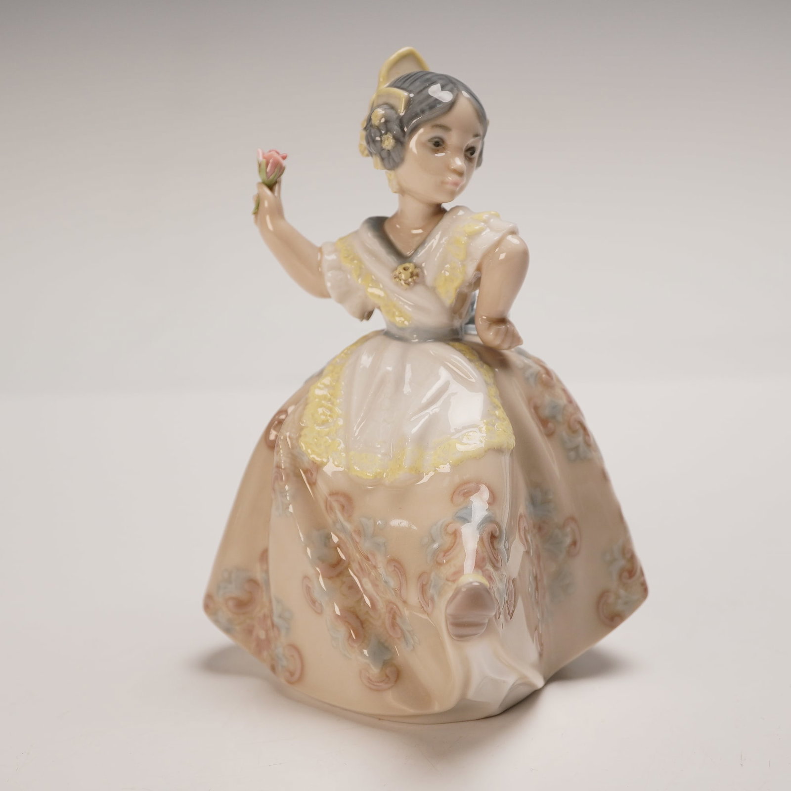 Lladro Porcelain Figurine, Teresita 1005375: Rich in cultural charm, this Lladro glazed porcelain figurine titled Teresita (model 1005375) depicts a young girl in traditional Spanish dress holding a single rose. Her gown is decorated with intric