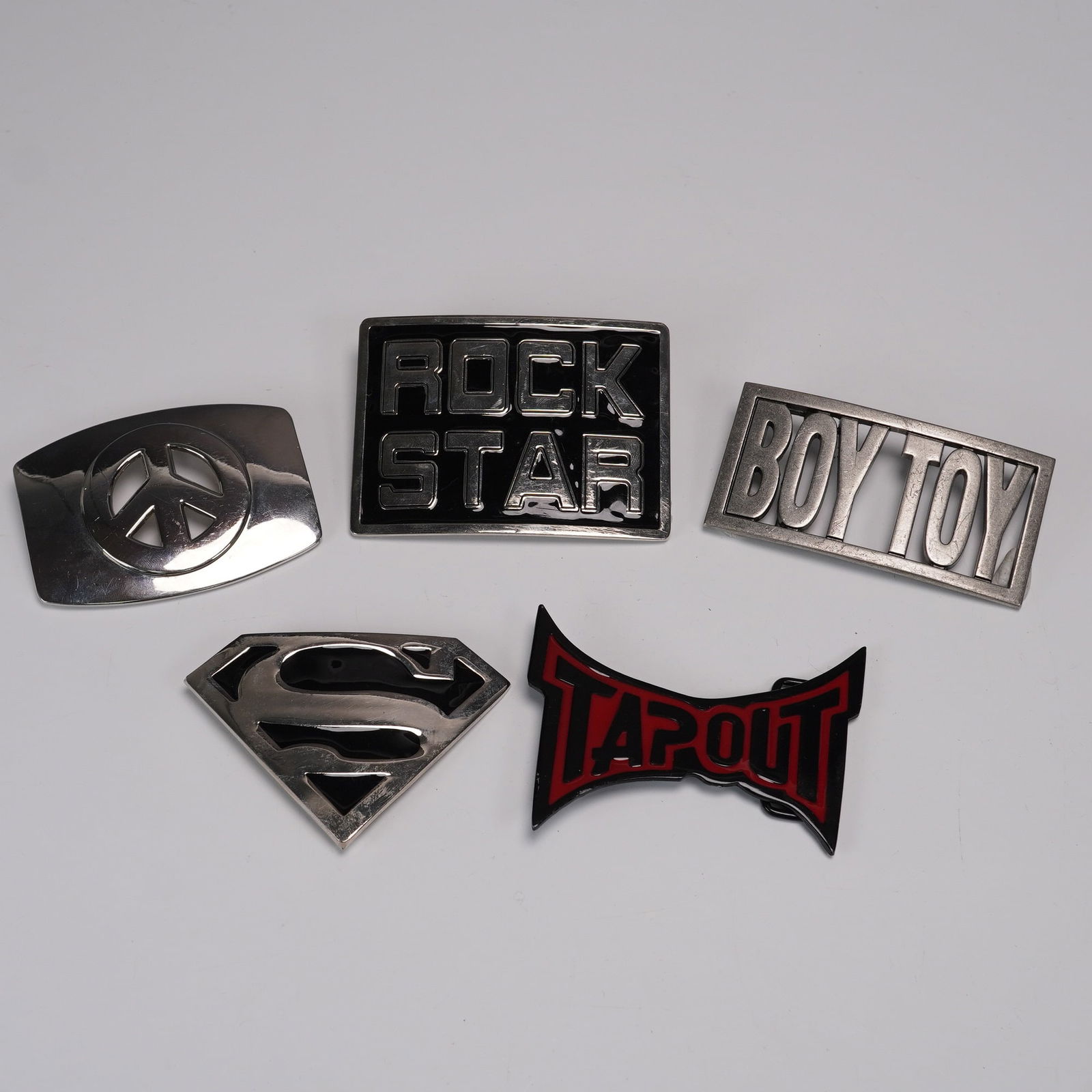 Five Pop Culture Belt Buckles Peace Tapout Superman: Set of five metal belt buckles featuring bold pop culture and icon designs including a raised peace sign, rectangular ROCK STAR block lettering, cutout BOY TOY text, Superman logo in polished silver t