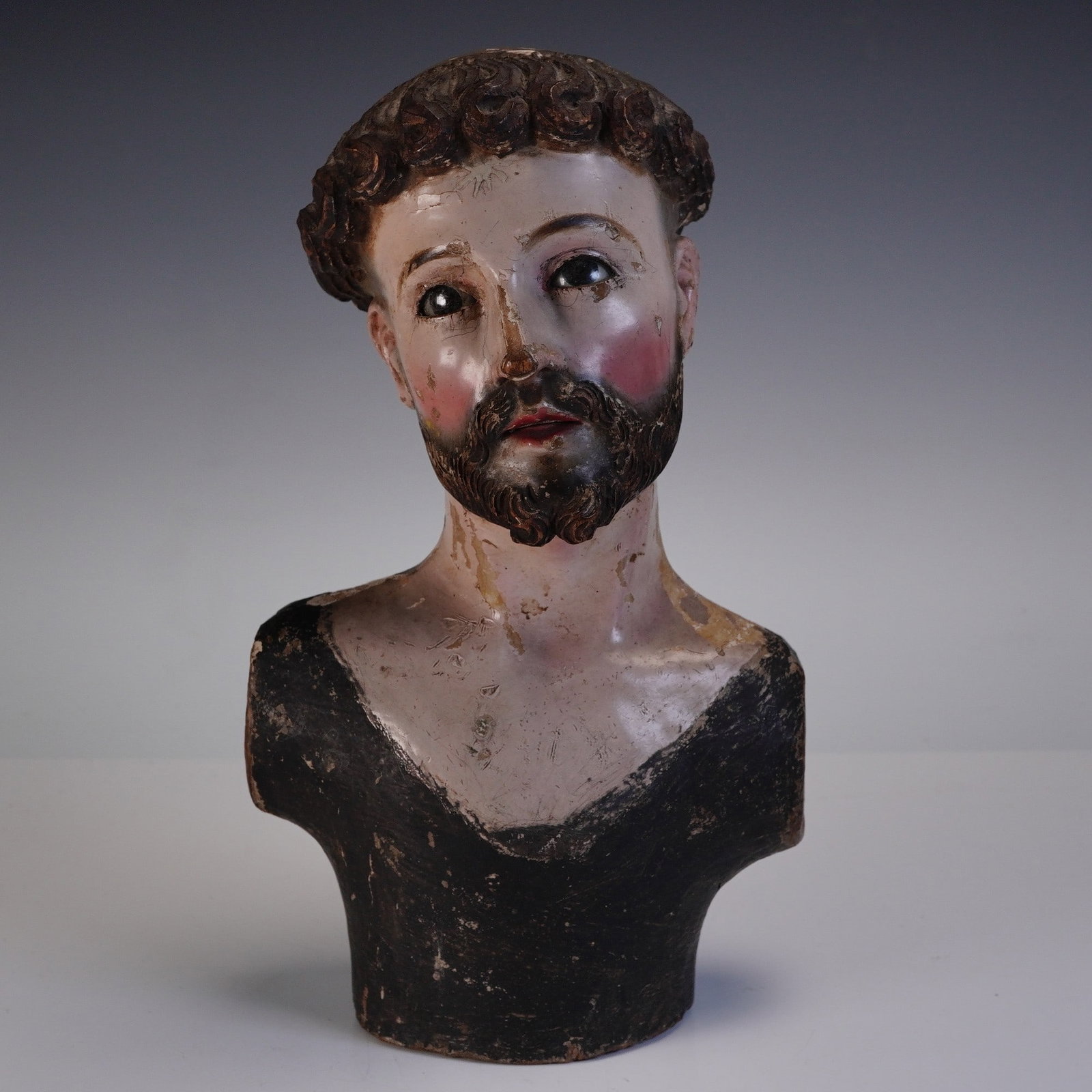 Wooden Santos Bust of a Male Saint with Glass Eyes: Bust of a male saint with glass brown eyes, featuring carved and painted details including a beard, mustache, and curled hair with a tonsure. Rendered from the shoulders up, the figure is executed in 