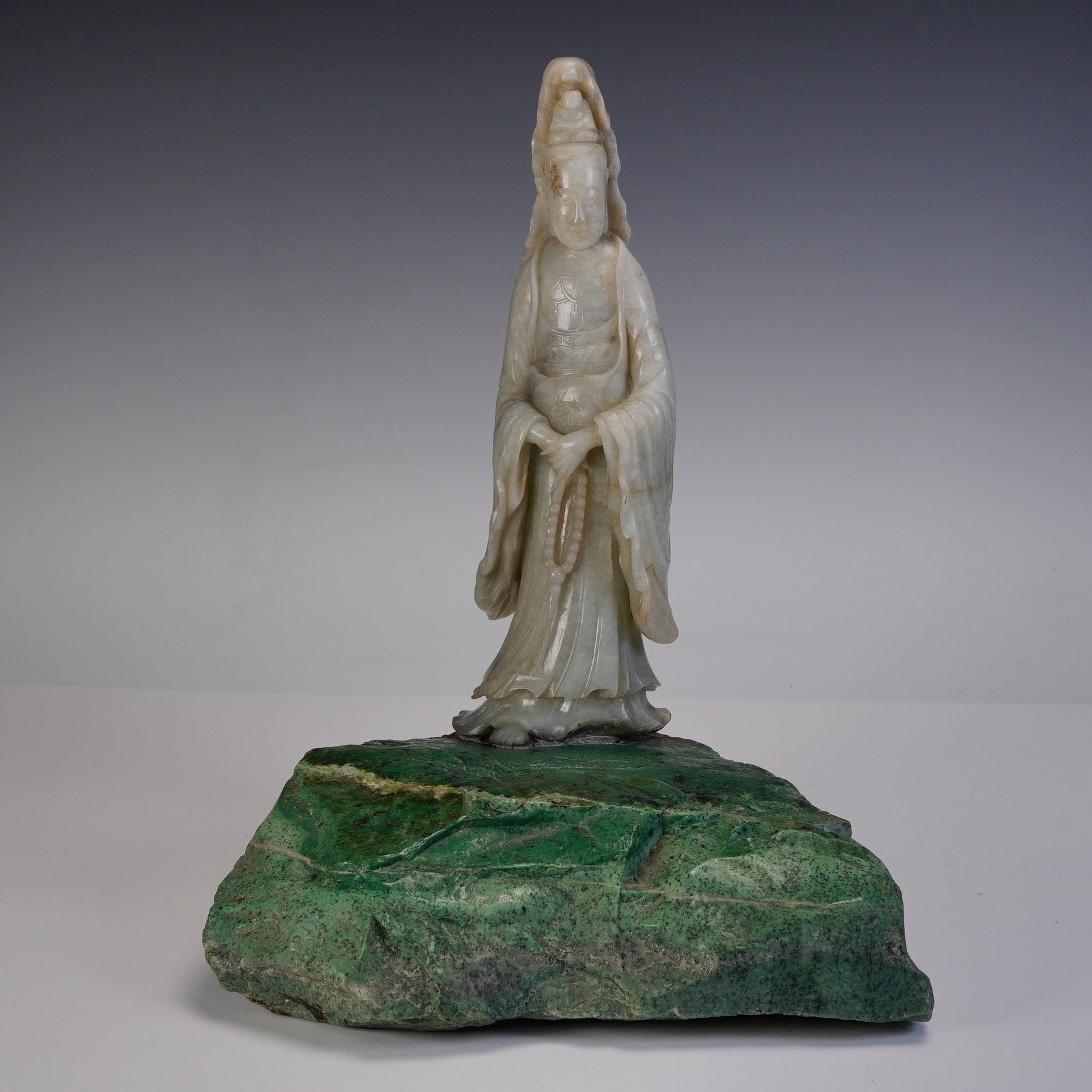 Chinese Jade Figure of Guanyin Mounted on Chloromelanite Base: Chinese carved jade figure depicting Guanyin, shown standing and holding a bead strand in both hands. The figure is carved from a grayish-white jade with natural russet and dark inclusions, detailed w