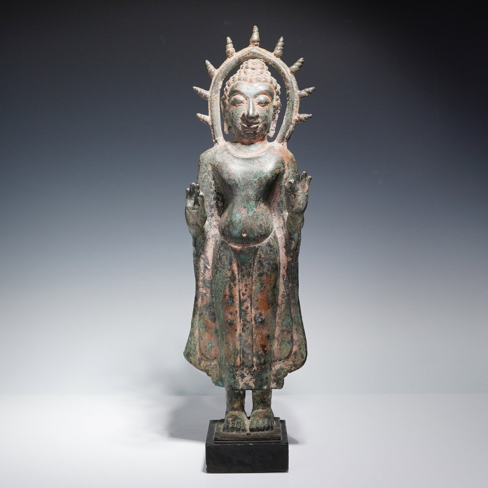 Thai Bronze Standing Buddha Sculpture: The figure is depicted in an upright posture with both hands raised in front, fingers positioned in a gesture. The head features a detailed ushnisha and stylized hair curls, with an arched halo behind