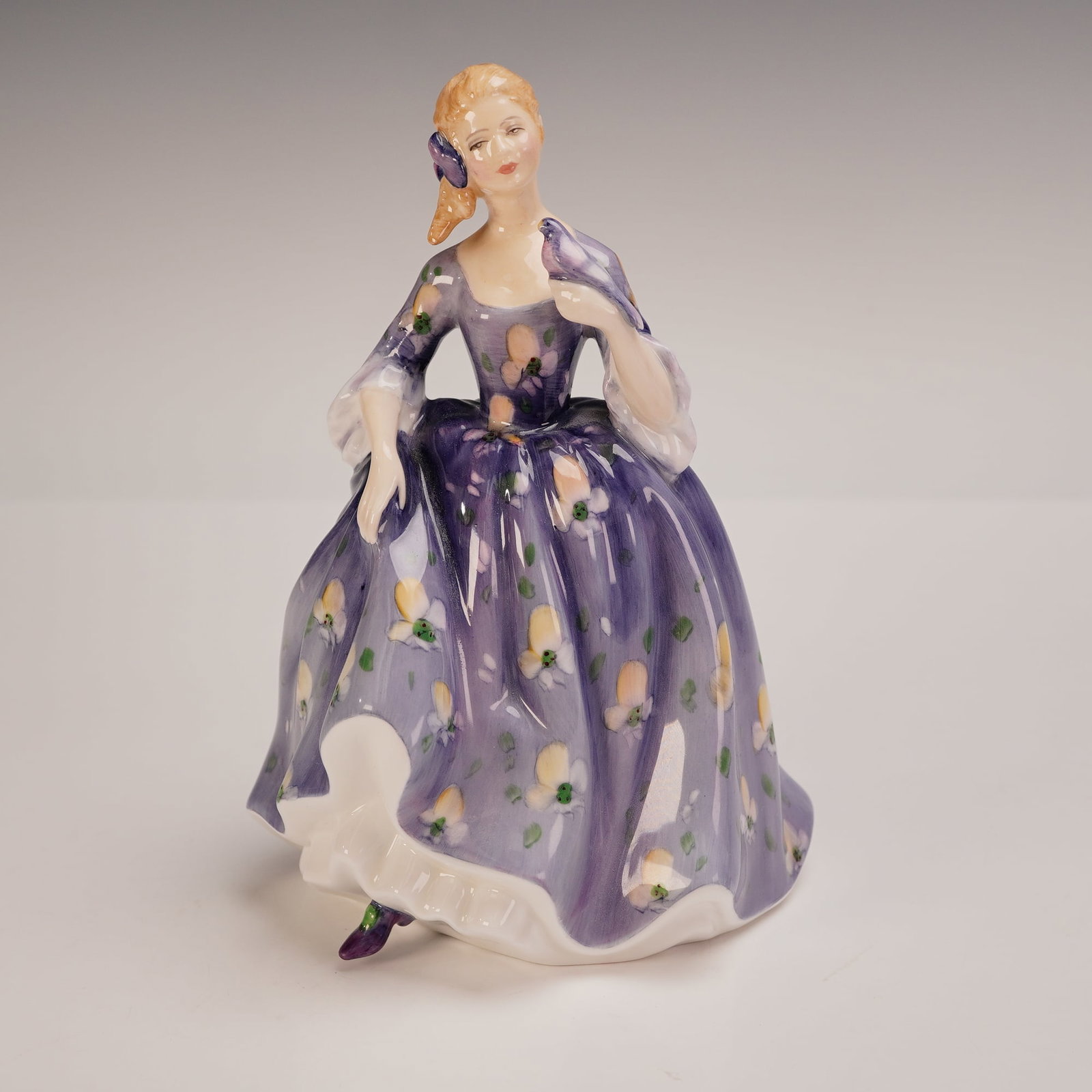 Royal Doulton Figurine Nicola HN2839: This charming Royal Doulton figurine titled Nicola HN2839 features a young woman in a flowing purple floral dress, with a purple ribbon in her hair and a matching bird perched on her shoulder. Designe