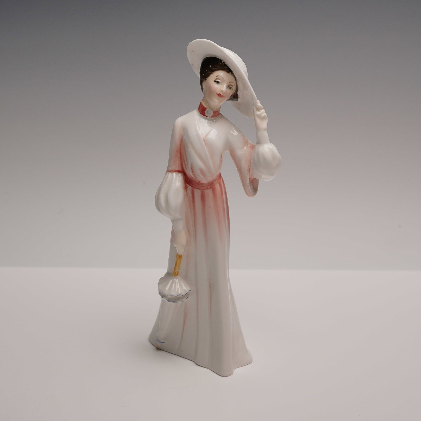 Royal Doulton Figurine, Catherine in Spring HN3006: A woman lifts her hand to her wide white hat, the brim traced with a soft blush to match the gentle sweep of rose over her flowing gown. Her robe glows with warm pink highlights that deepen around her