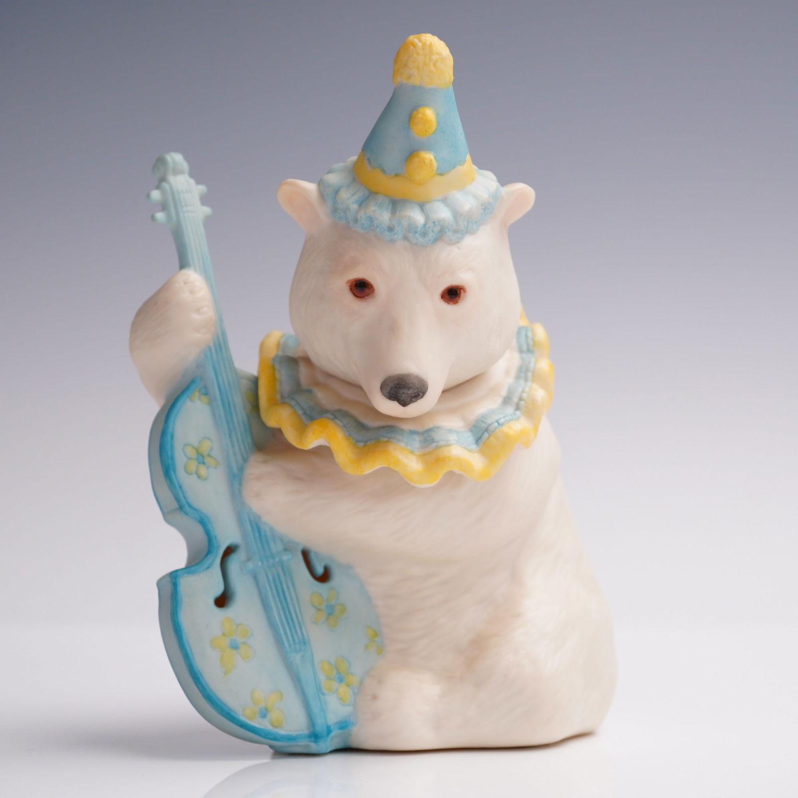Cybis Porcelain Figurine, Polar Bear Musician: Charming Cybis porcelain figurine depicting a whimsical polar bear dressed as a clown, seated while playing a pastel blue upright bass adorned with hand-painted yellow flowers. The bear features a scu