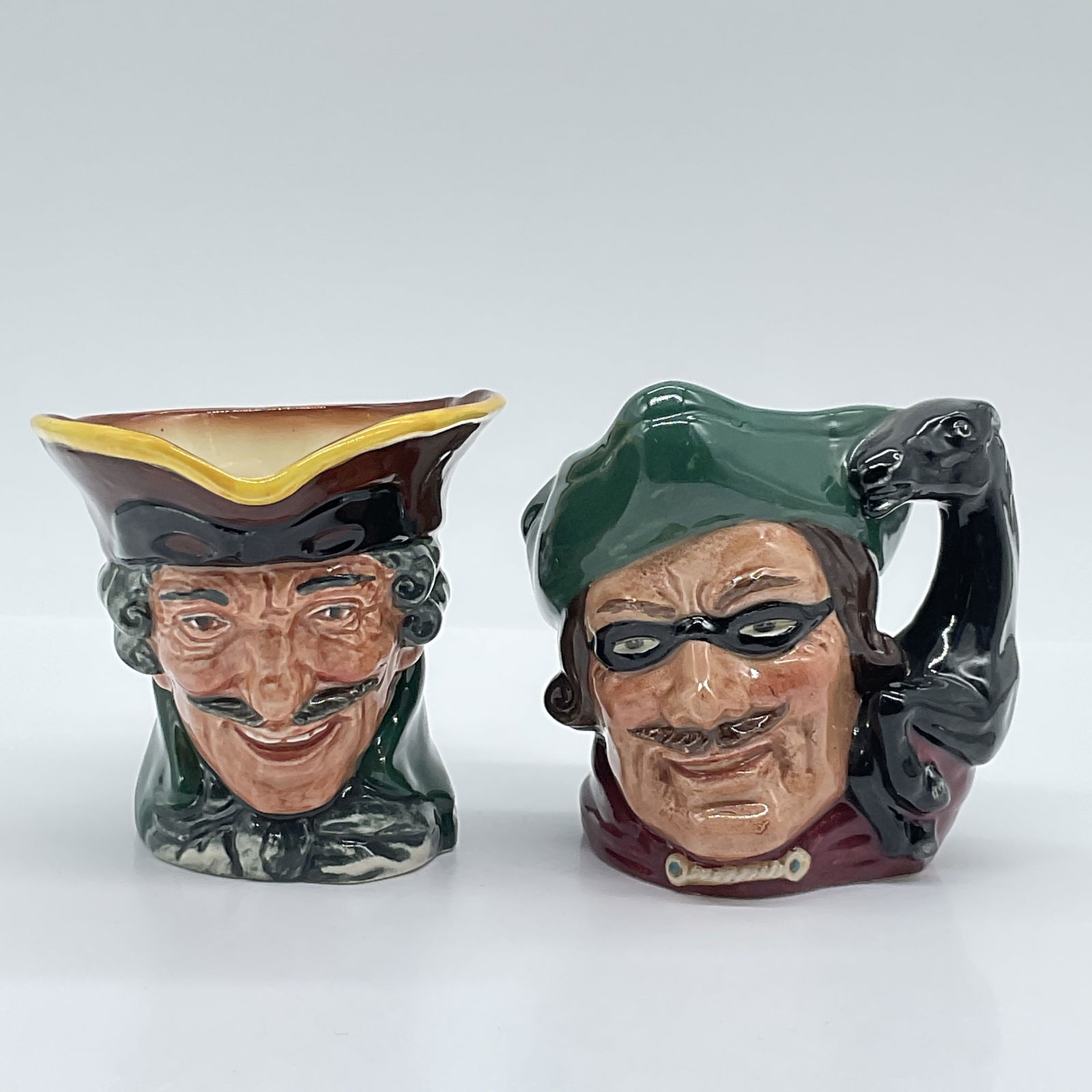 2pc Royal Doulton Character Jugs, Dick Turpin D6535 & D5618: Two Royal Doulton character jugs featuring the infamous English highwayman, Dick Turpin. This lot includes the D6535 jug, designed by David B. Biggs, with a tri-corner hat and a pistol-shaped handle,