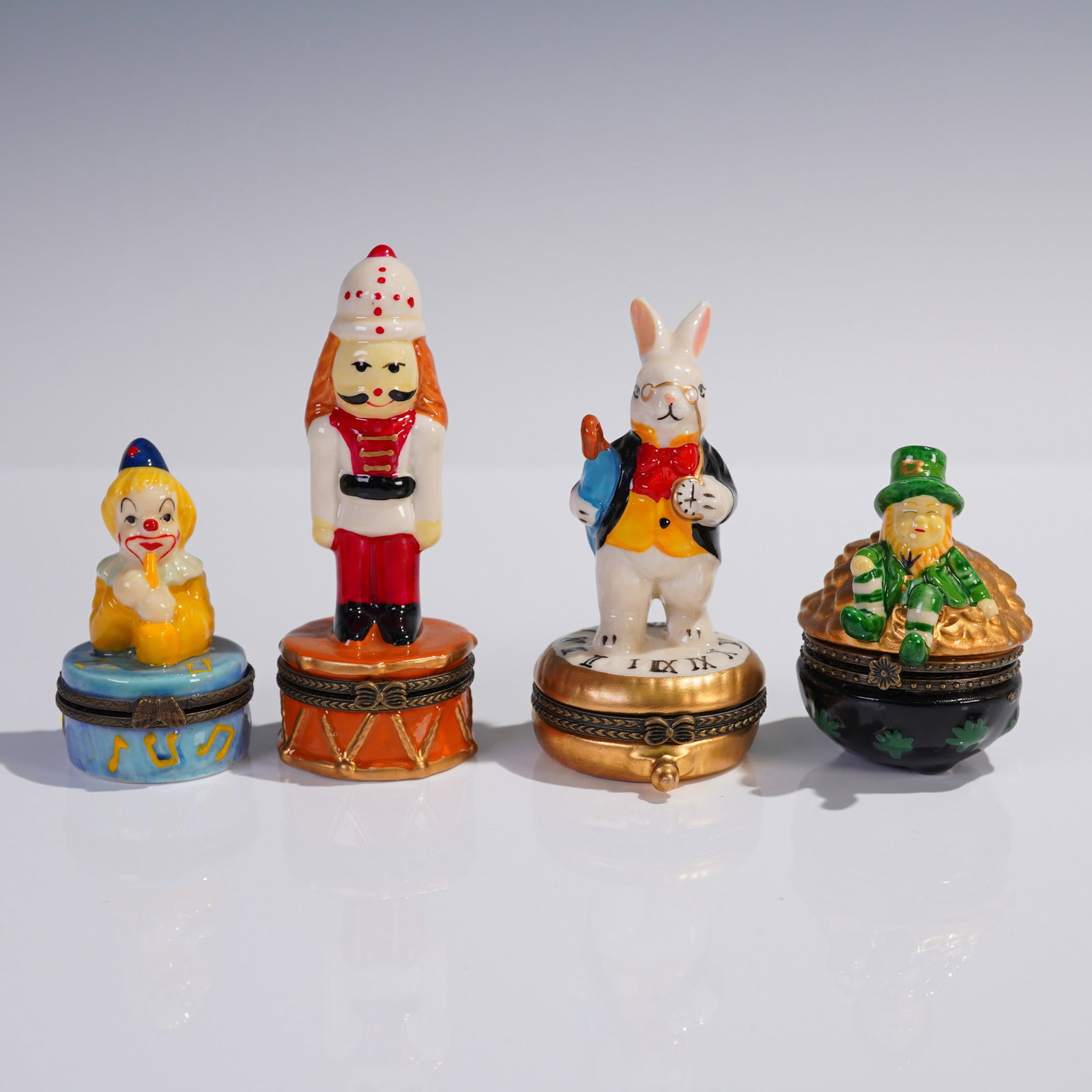 4pc Whimsical Figural Porcelain Trinket Box Quartet: 4-piece figural porcelain trinket box lot featuring a whimsical assortment of unmarked miniature hinged boxes. Includes a musical clown seated atop a blue drum, a nutcracker soldier standing on an ora