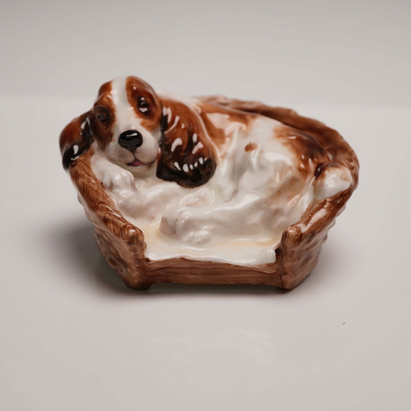 Royal Doulton Figurine, Cocker Spaniel Lying in Basket HN2585: Model no. 1155; depicts a dog resting in his basket. Modeled in white with brown and black markings; light brown basket. Hand written HN number. Royal Doulton backstamp. Issued: 1941 - 1985 Dimensions