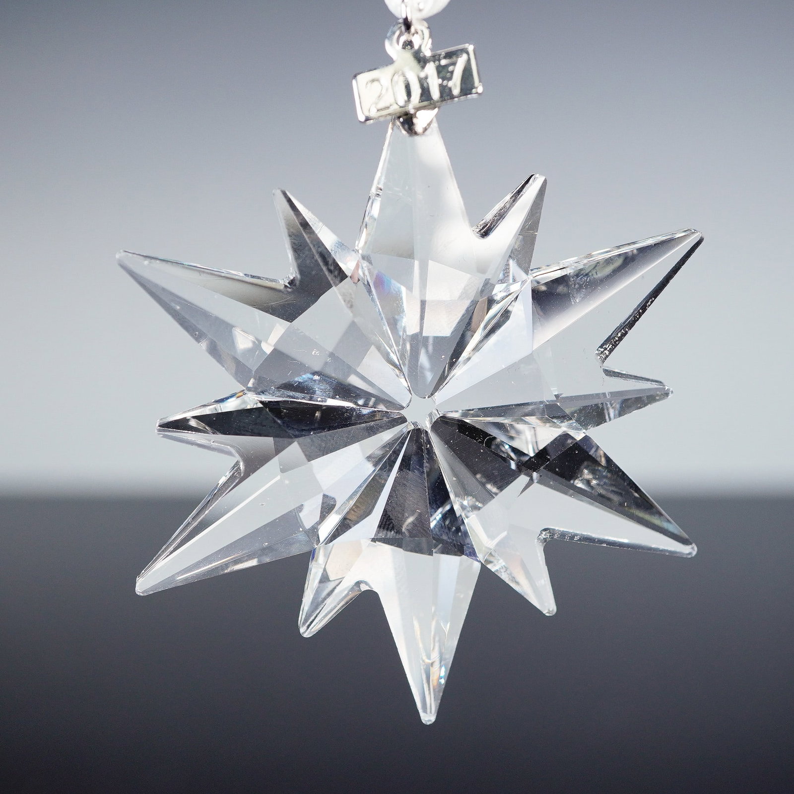 Swarovski Crystal Tree Ornament, Star 2017 5257592: This clear faceted crystal ornament is designed in the form of a twelve-pointed star with intricate cuts that reflect and refract light. It is suspended from a white satin ribbon and includes a polish