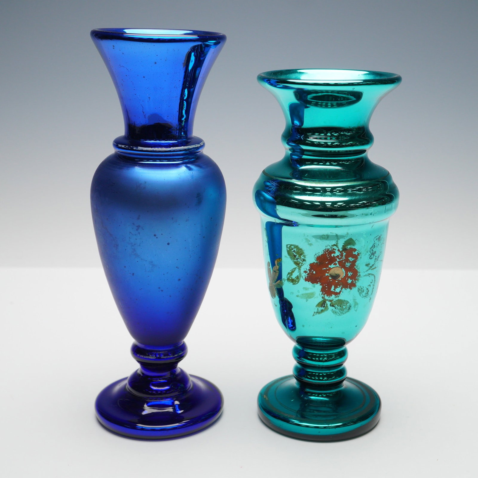 Pair of Mercury Glass Cobalt & Teal Floral Vases: A duo of pedestal based mercury glass vases, including one in a deep cobalt blue and a vivid teal with a lustrous mirrored surface and hand painted floral embellishment in red, green and gold tones. T
