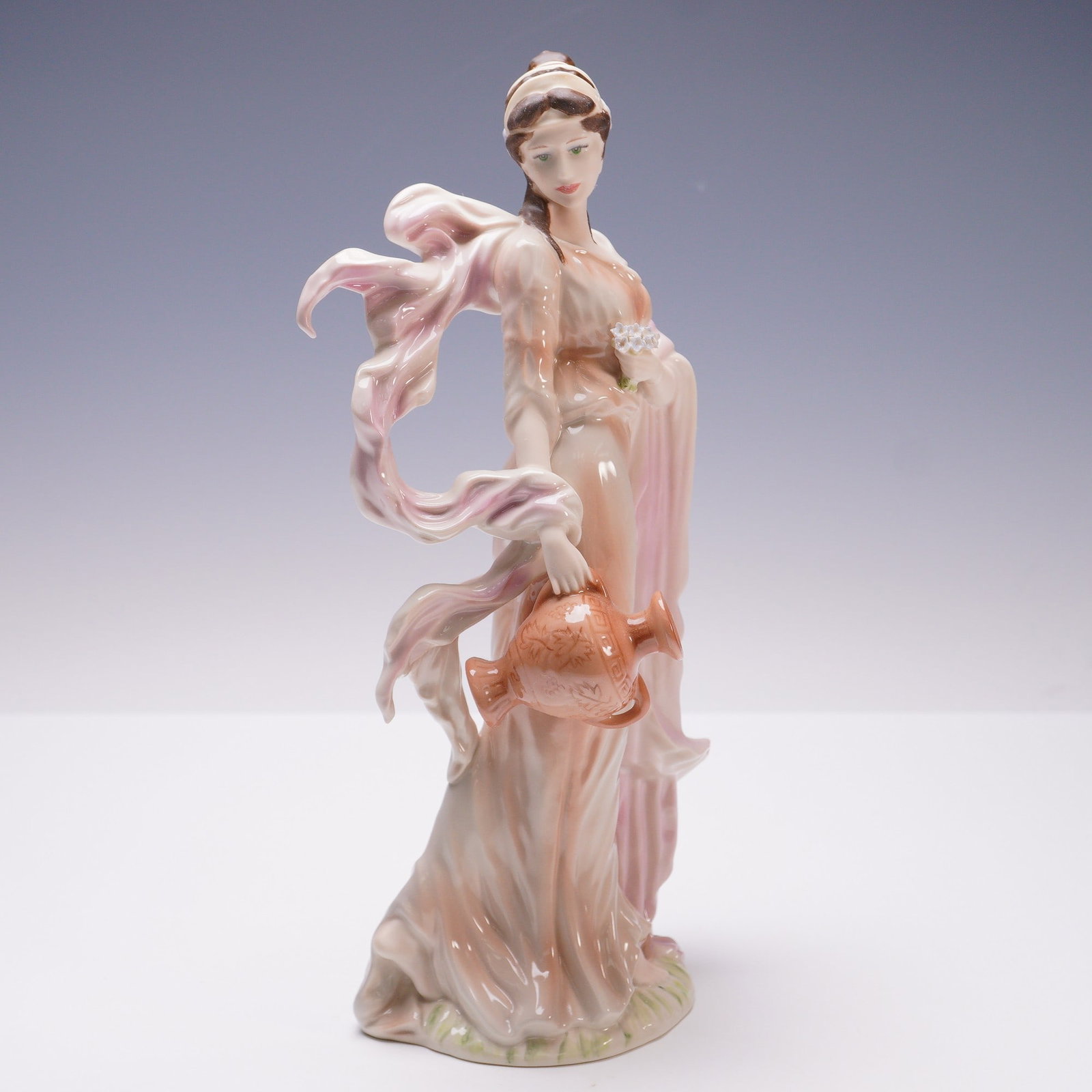 Wedgwood Porcelain Figurine, Winsome: Glazed finish porcelain figure from The Classical Collection. Portrays a serene woman clad in a soft, peach-toned gown, her drapery cascading gracefully around her and adorned with a windswept sash th