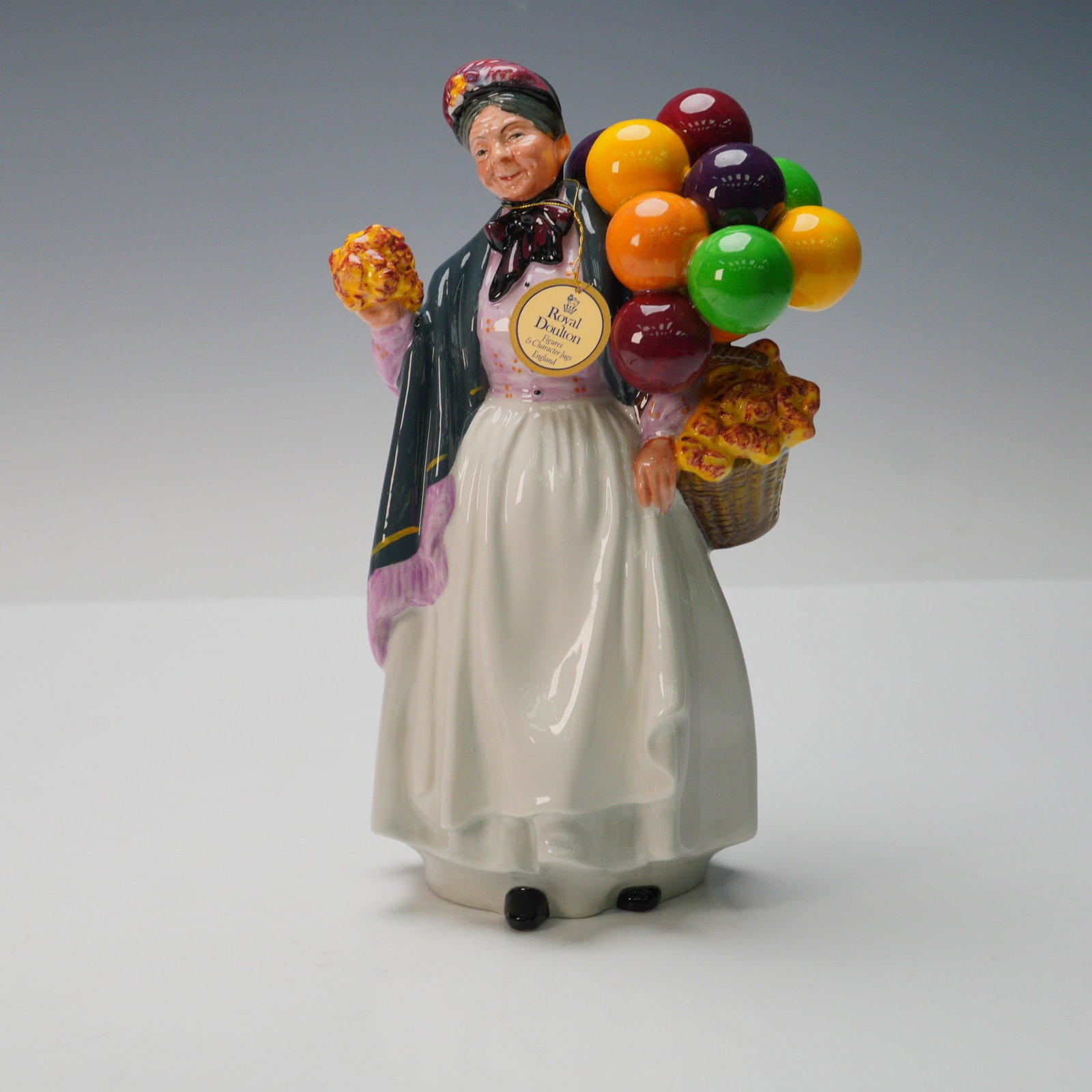 Royal Doulton Figurine, Biddy Penny Farthing HN1843: From the Balloon Character series. She is dressed in long dress with grey and lavender shawl carrying multicolored balloons and flowers. Royal Doulton hang tag. Royal Doulton backstamp. Artist: Leslie
