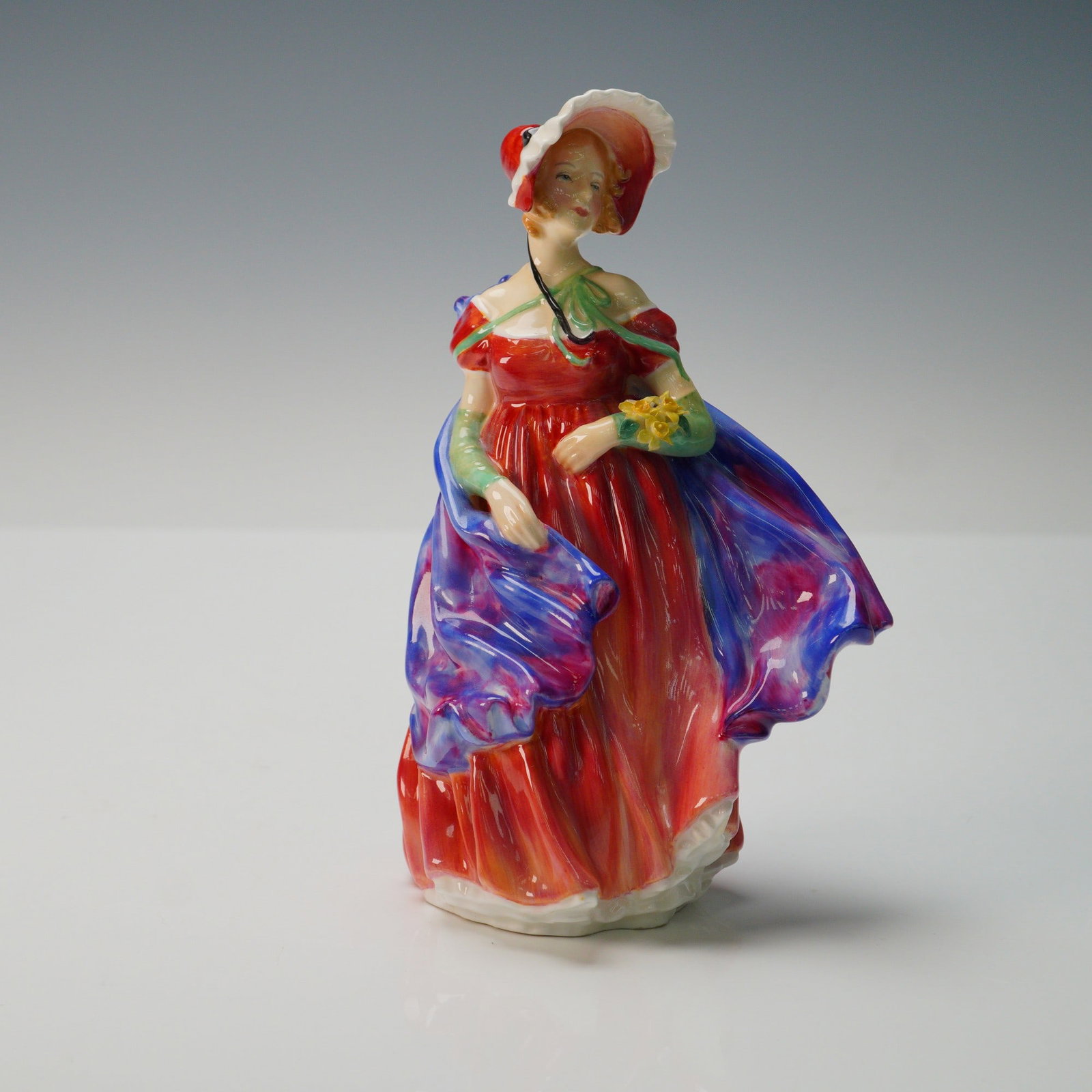 Royal Doulton Figurine, Lady April HN1958: A Royal Doulton bone china figurine titled Lady April HN1958, modelled as a graceful young woman in a flowing red gown with green gloves, holding a small bouquet of yellow flowers, with a vibrant purp