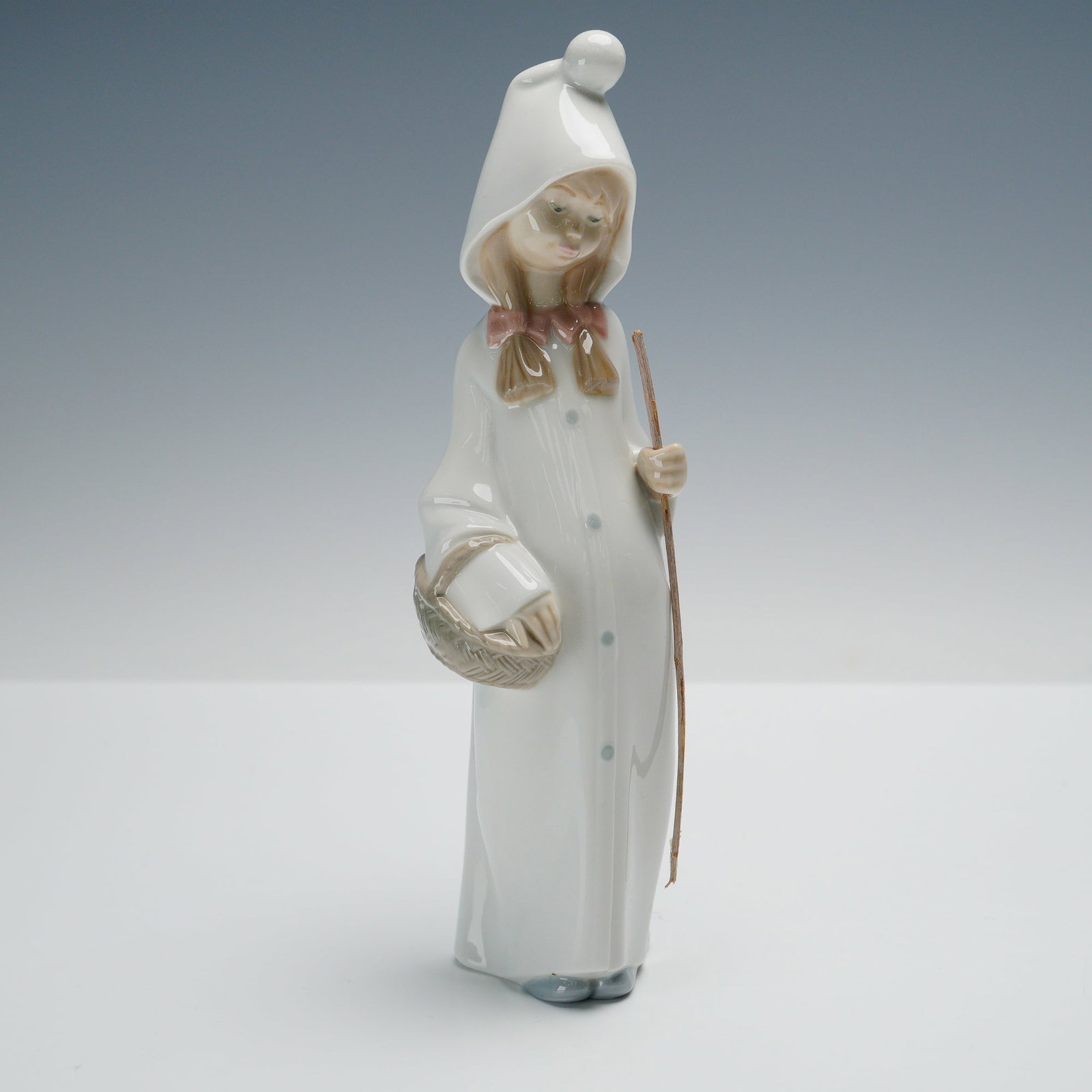 Lladro Porcelain Figurine, Girl with Basket 1004678: Glossy finish porcelain depicting a young girl in a long white hooded cloak, the hood tipped with a pom, carrying a slender wooden staff in one hand and a woven basket in the other. The figure is rend