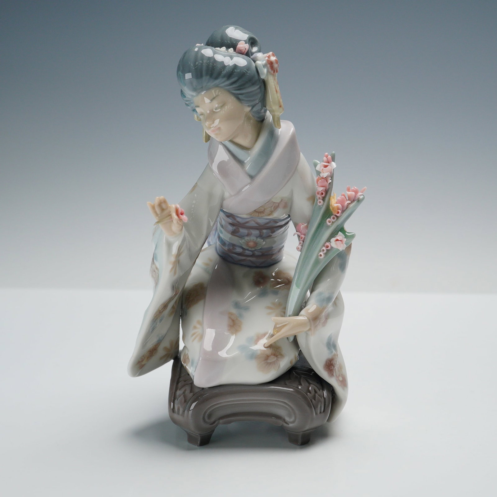 Lladro Porcelain Figurine, Kiyoko 1001450: Glossy finish depicting a finely detailed geisha in a flowing kimono adorned with delicate floral patterns, her hair styled in an elaborate coiffure accented with blossoms. She kneels gracefully on a 