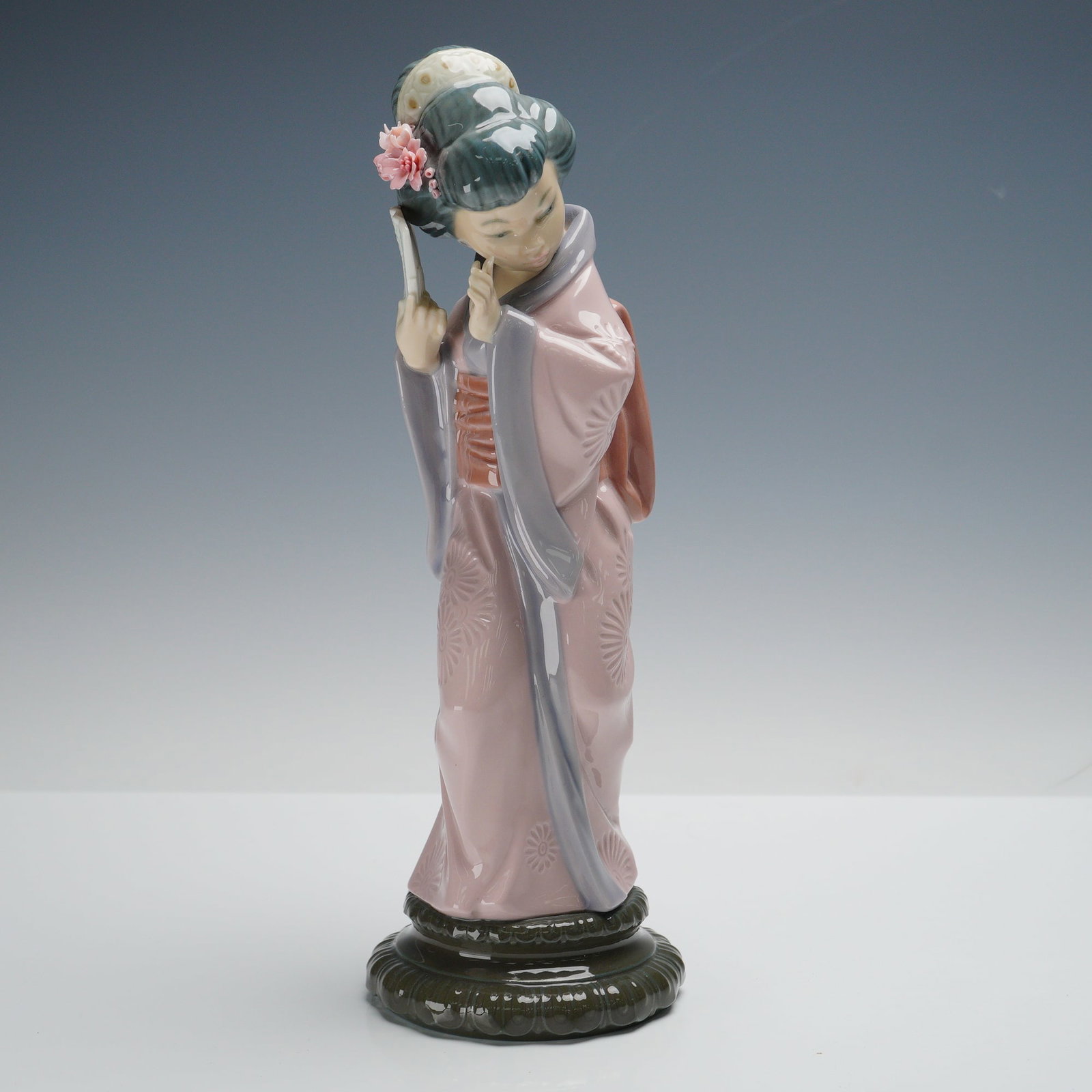 Lladro Porcelain Figurine, Chrysanthemum 1004990: A Lladro porcelain figurine titled Chrysanthemum 1004990, depicting a graceful geisha in a pale pink kimono with floral patterns, holding a folding fan and adorned with a flower in her hair. The figur