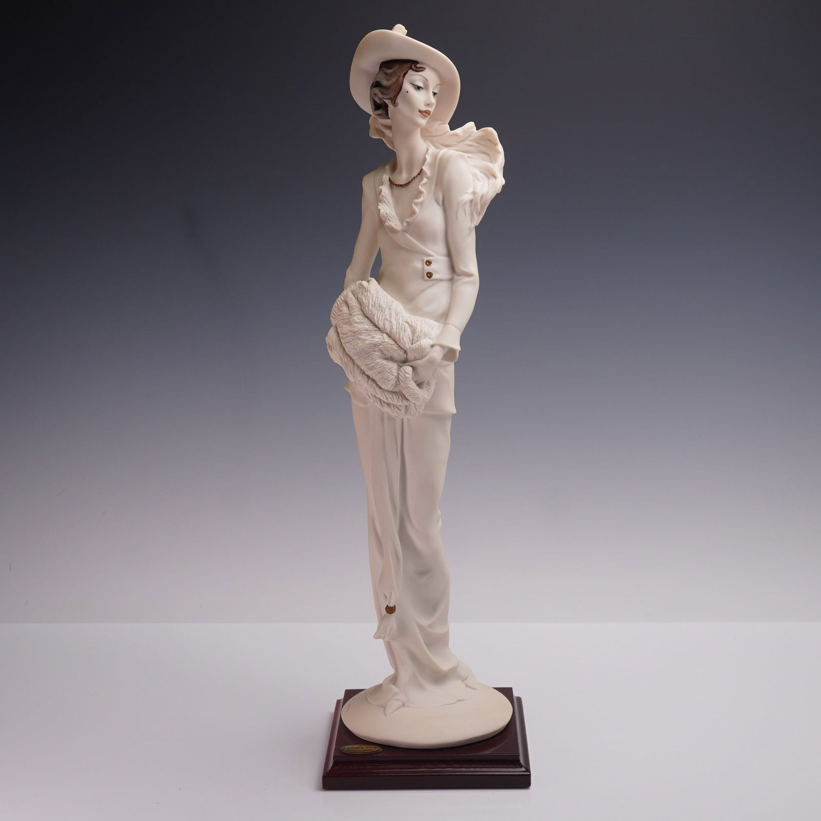 Florence Giuseppe Armani Figurine of a Lady: A porcelain figurine depicting a woman in period attire with a wide-brimmed hat, ruffled collar, and fur muff. Features artists signature and manufacturers marks included. Mounted on a wooden base wit