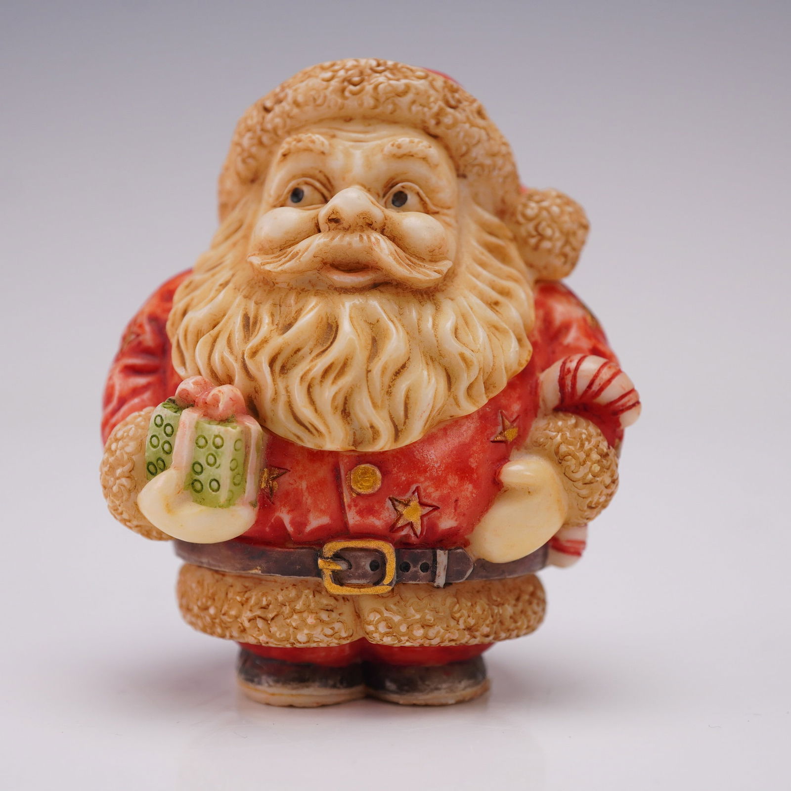 Pot Bellys Oddbods Covered Box Figurine, Santa With Present: The figure depicts Santa Claus in a red coat decorated with gold stars, holding a green present in one hand and a candy cane in the other. The removable head reveals a hidden compartment containing a