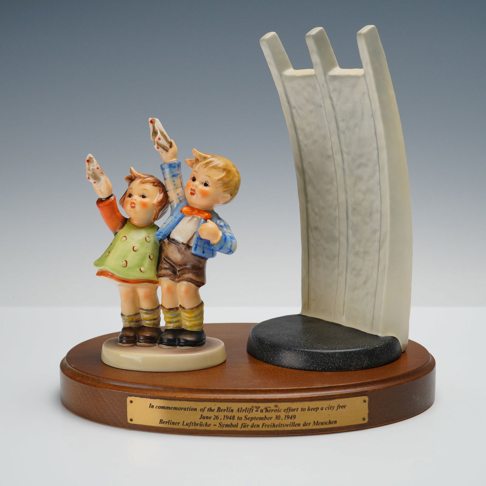 3pc Goebel Hummel Berlin Airlift Commemorative Figurine: Goebel Hummel Berlin Airlift commemorative figurine group featuring two children waving, accompanied by a sculptural representation of the Berlin Airlift Memorial, mounted on a wooden plinth with engr