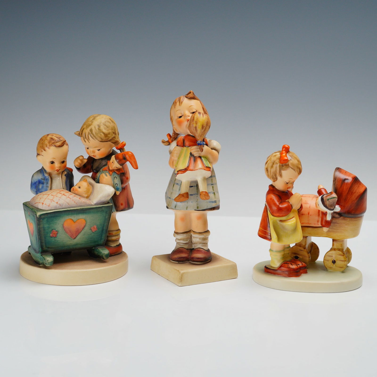 3pc Goebel Hummel Figurines: Figurines including 'Kiss Me' (311) depicting a little girl with her doll, 'Blessed Event' (333) featuring siblings with a newborn baby, and 'Doll Mother' (67) showing a little girl with a doll in a b