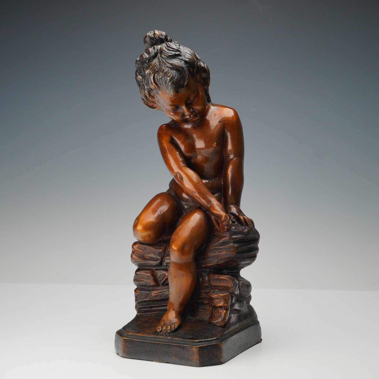 Bronzed Figural Sculpture of a Seated Girl: Cast sculpture depicting a young girl seated on a rocky outcrop, her head gently inclined and arms resting on her lap in a serene pose. The figure is modeled with smooth contours and softly detailed h