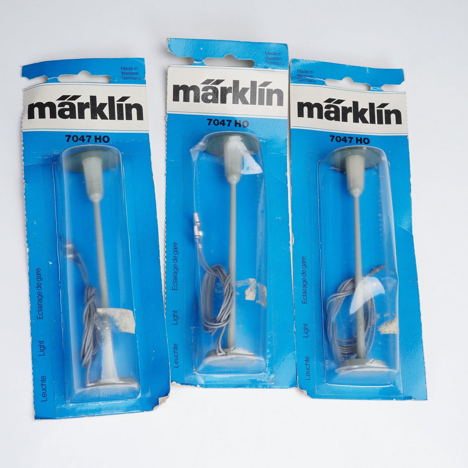 3pc Marklin Model Street Lamp Set, Light 7047HO: Three-piece set, is presented in its original blue retail packaging marked and labeled Leuchte Light Eclairage de gare. The lamps feature a tall white post with a round base, wiring attached, and a mo
