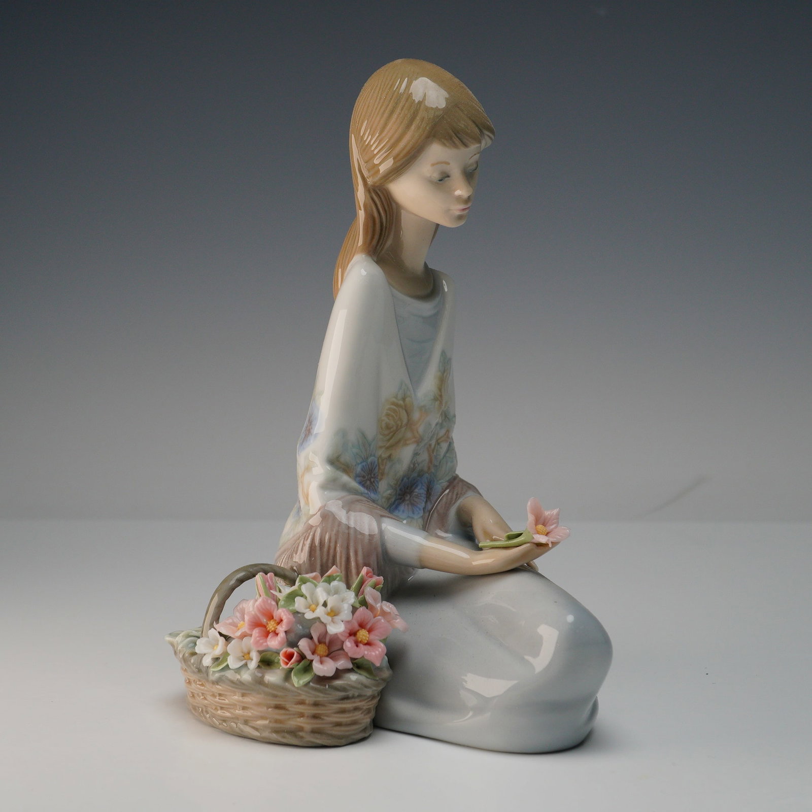 Lladro Porcelain Figurine, Flower Song 1007607: Lladro porcelain figurine titled Flower Song number 1007607 depicting a young girl in a pastel floral dress seated with a basket of delicate hand-crafted flowers and holding a single bloom. The figuri